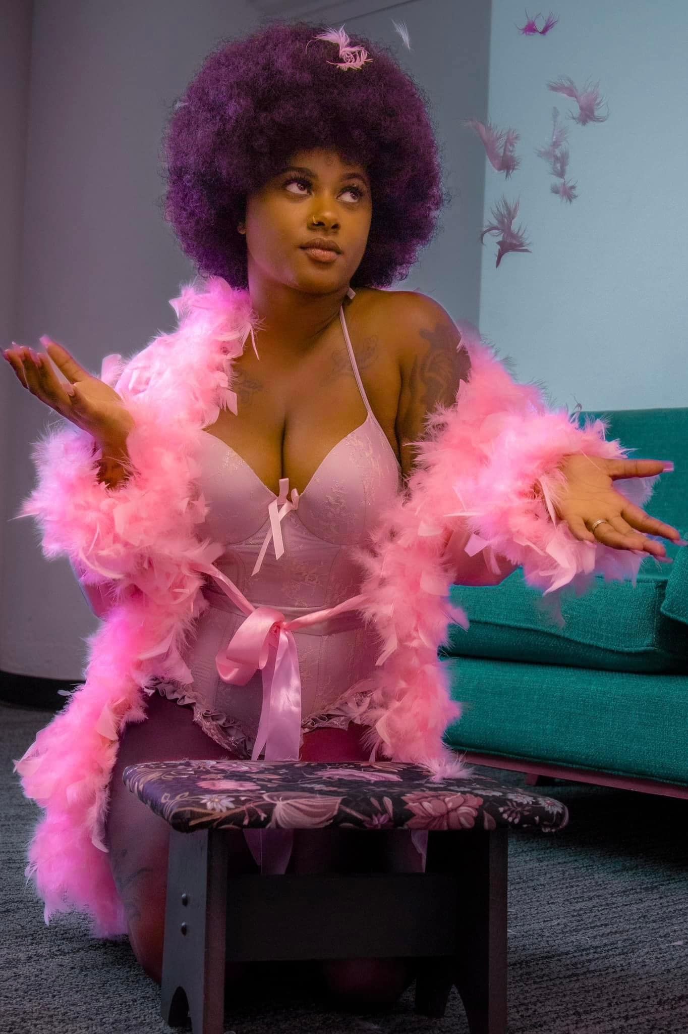 Woman with purple afro wearing pink feather boa and lingerie posing on a stool indoors.