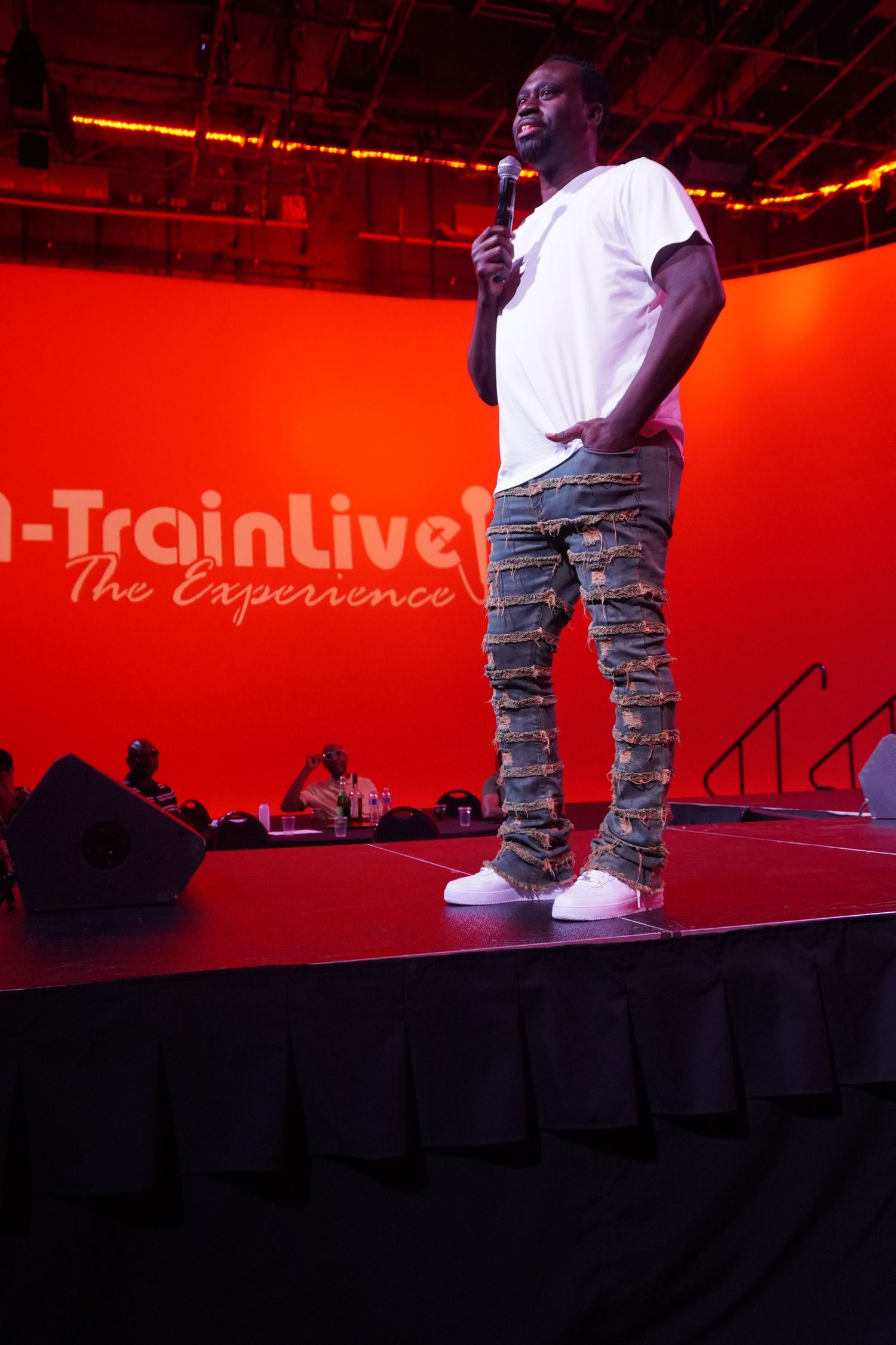 A performer in white tee and camo pants holds mic on stage at A-Train Live The Experience.