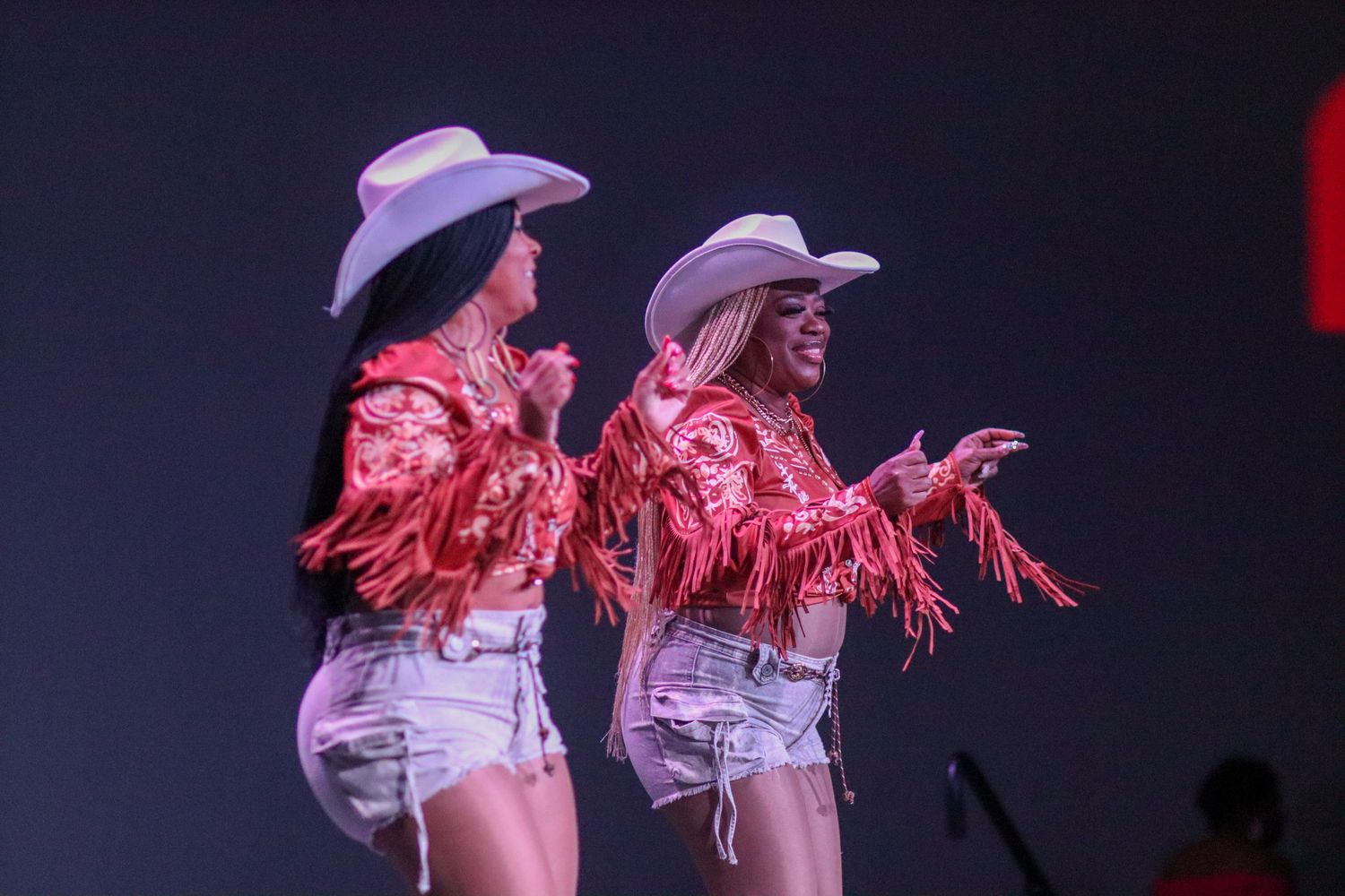 Two women in red fringe jackets and cowboy hats perform on a lit stage.