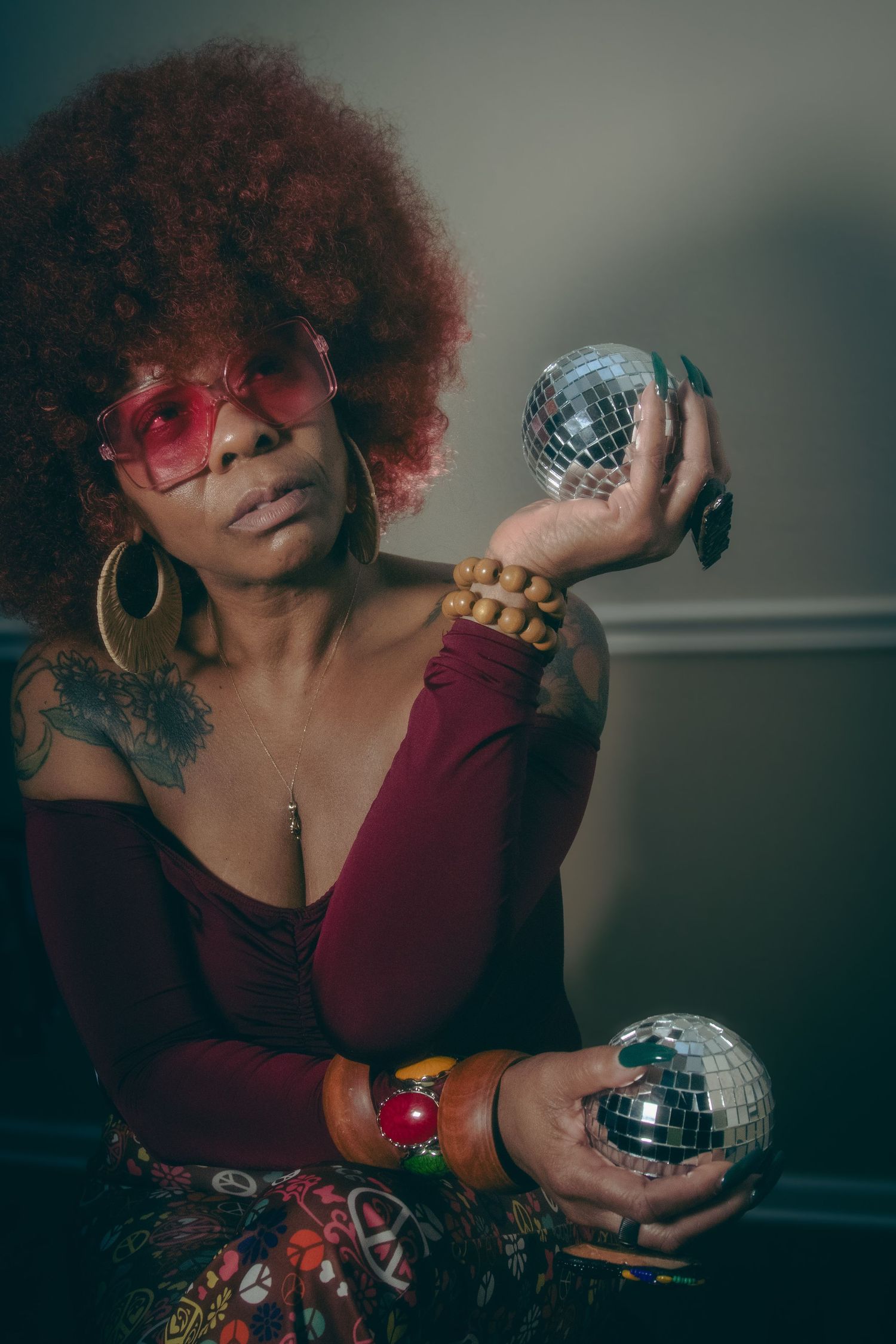 Woman with red afro and sunglasses holding two disco balls in a retro-inspired studio portrait.