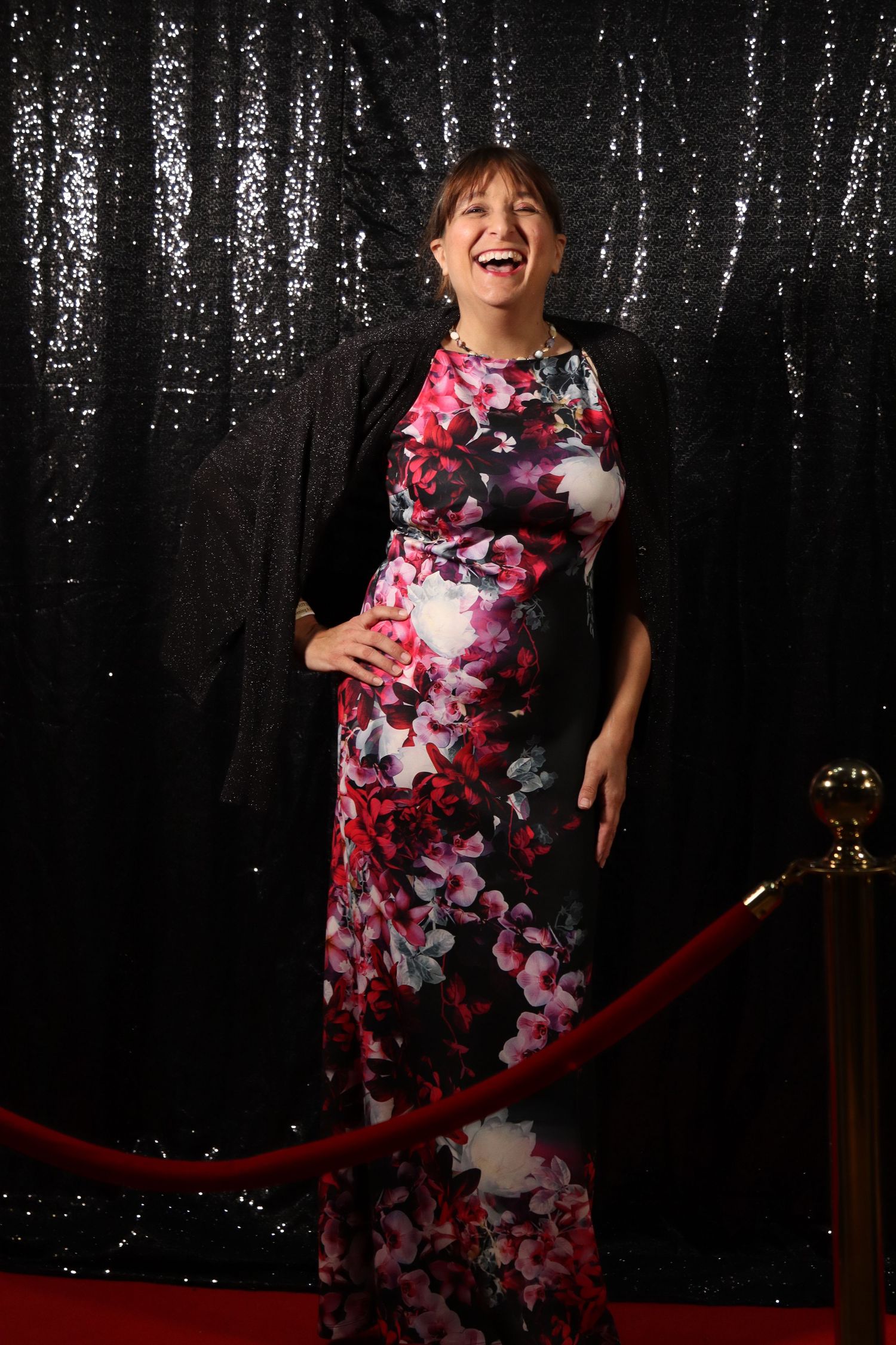 Woman laughing in floral gown on red carpet with sequin backdrop at formal event.