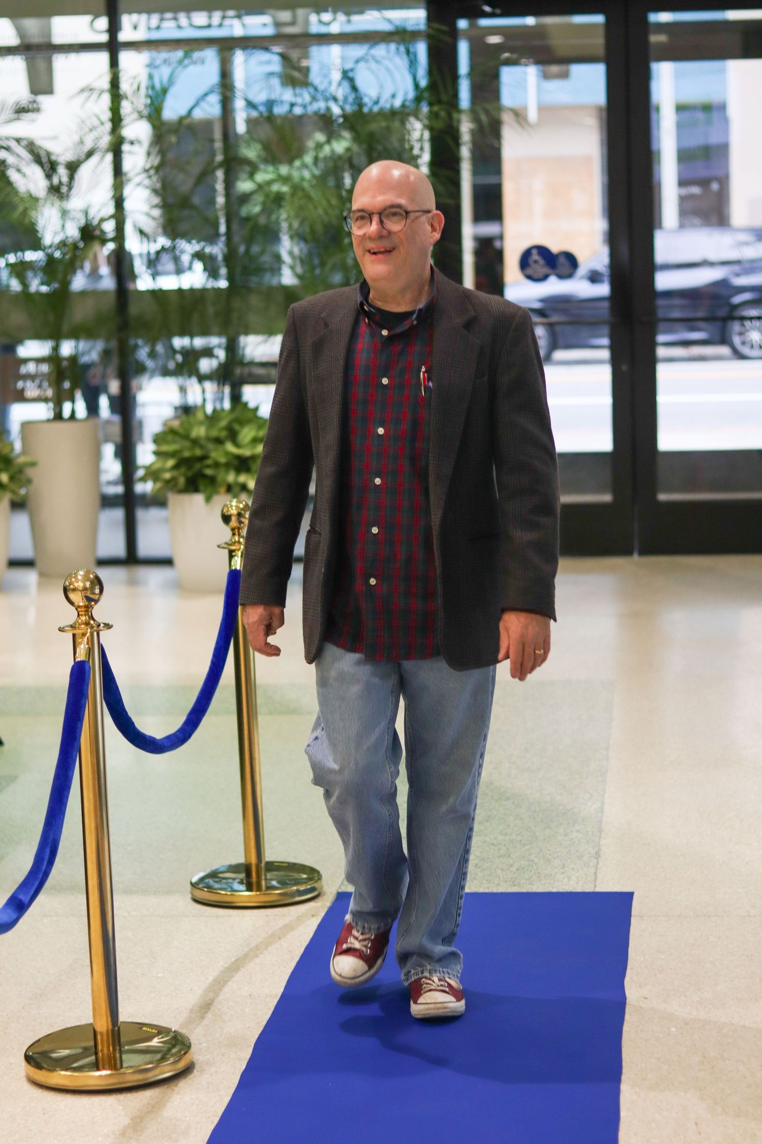 Smiling bald man in blazer and plaid shirt walks blue carpet at formal event entrance.