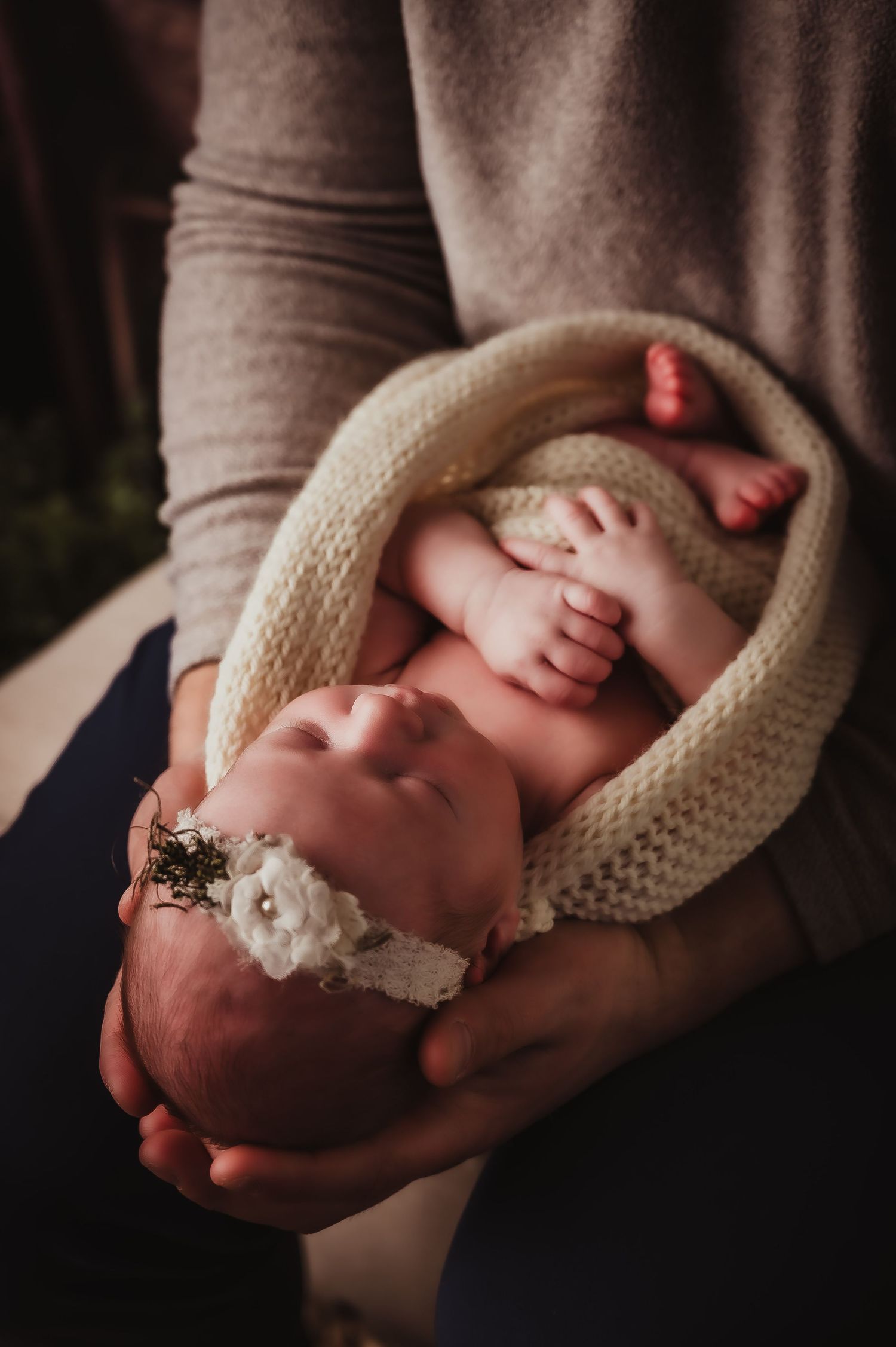 Newborn in knit wrap wearing delicate headband nestled in loving arms.