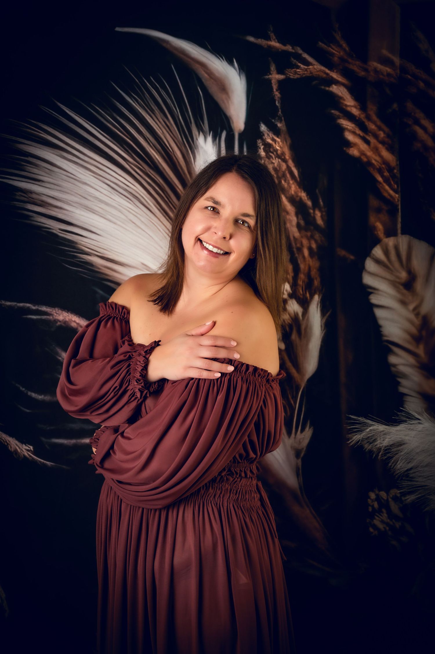 Portrait in burgundy off-shoulder dress against dramatic black background with white feathers.
