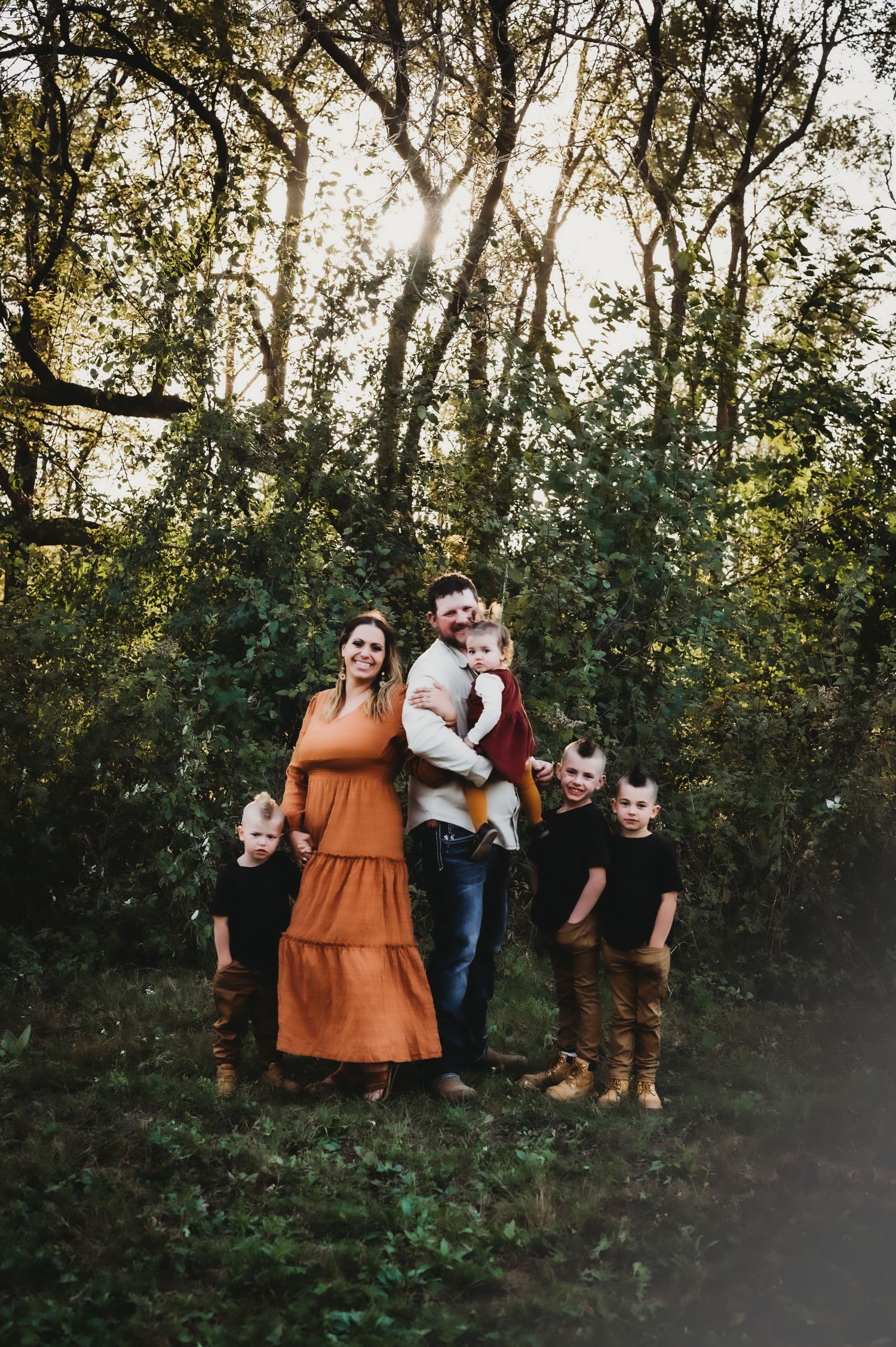 A family poses together in a wooded area at sunset with sunlight filtering through autumn trees in the background.