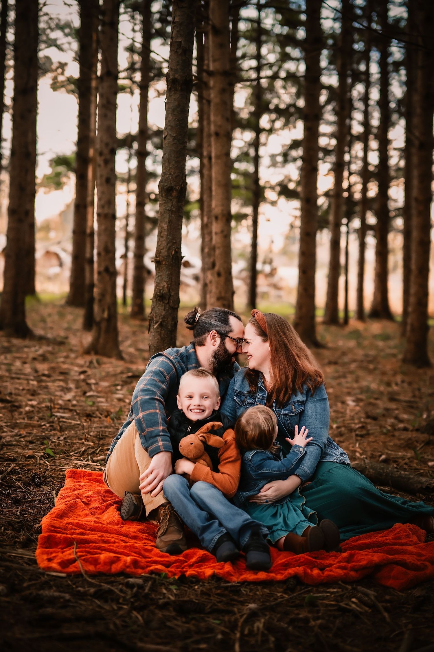 Family of four sitting on orange blanket in pine forest, parents kissing while children play with stuffed animal toy.