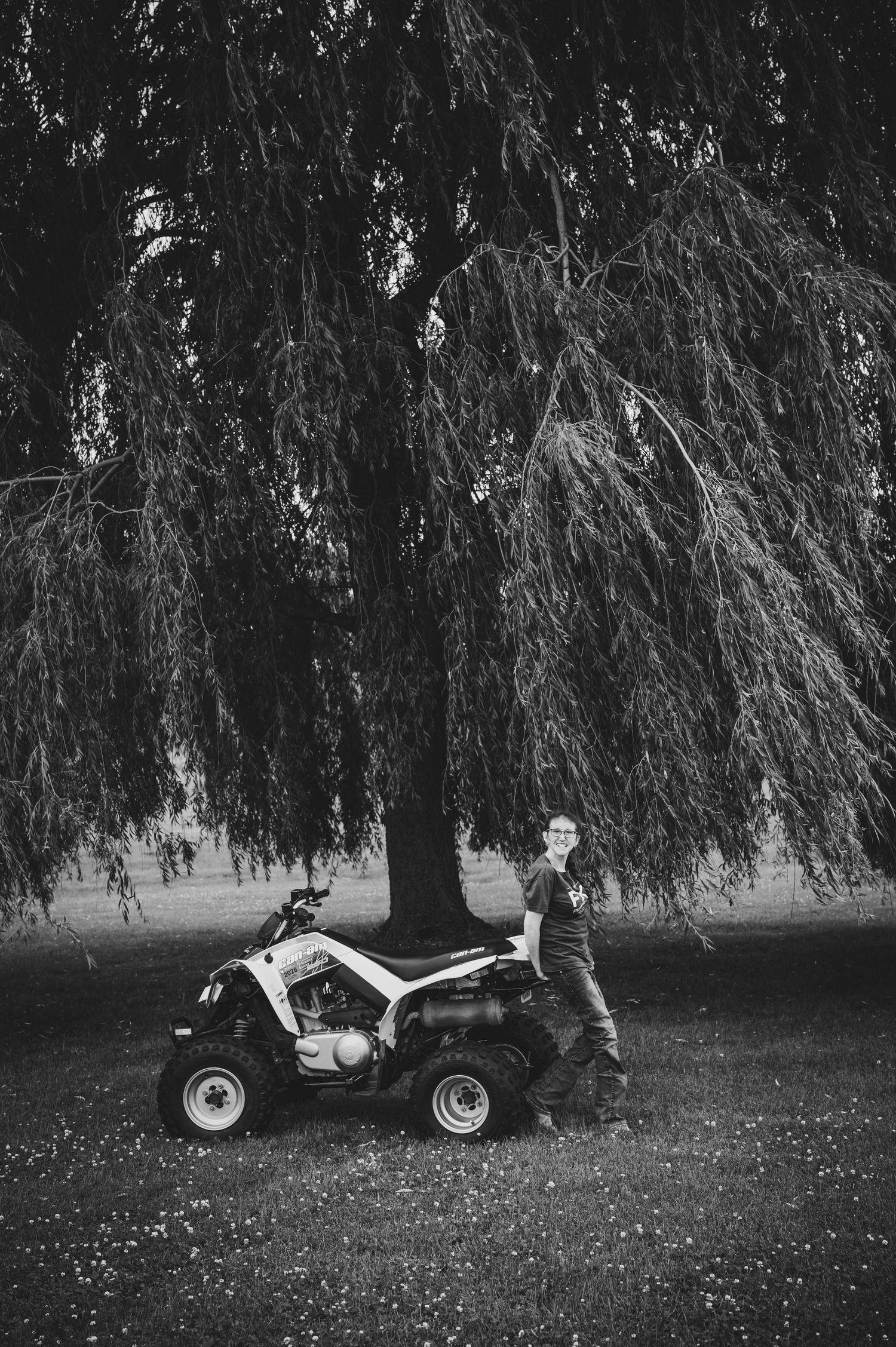 Black and white photo of a person standing beside an ATV quad bike under a large weeping willow tree on grass.