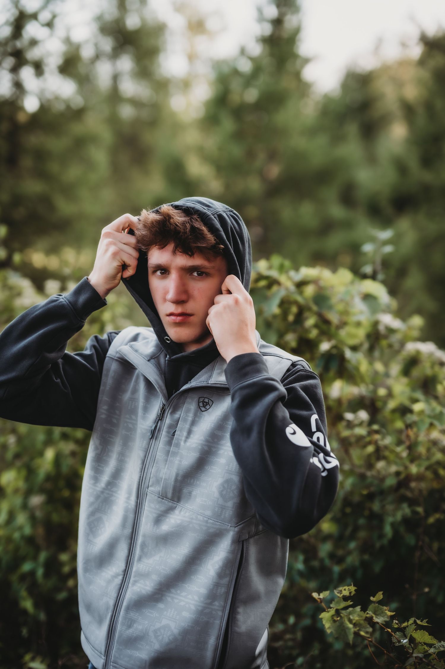 Person adjusting grey hoodie while standing among green foliage.