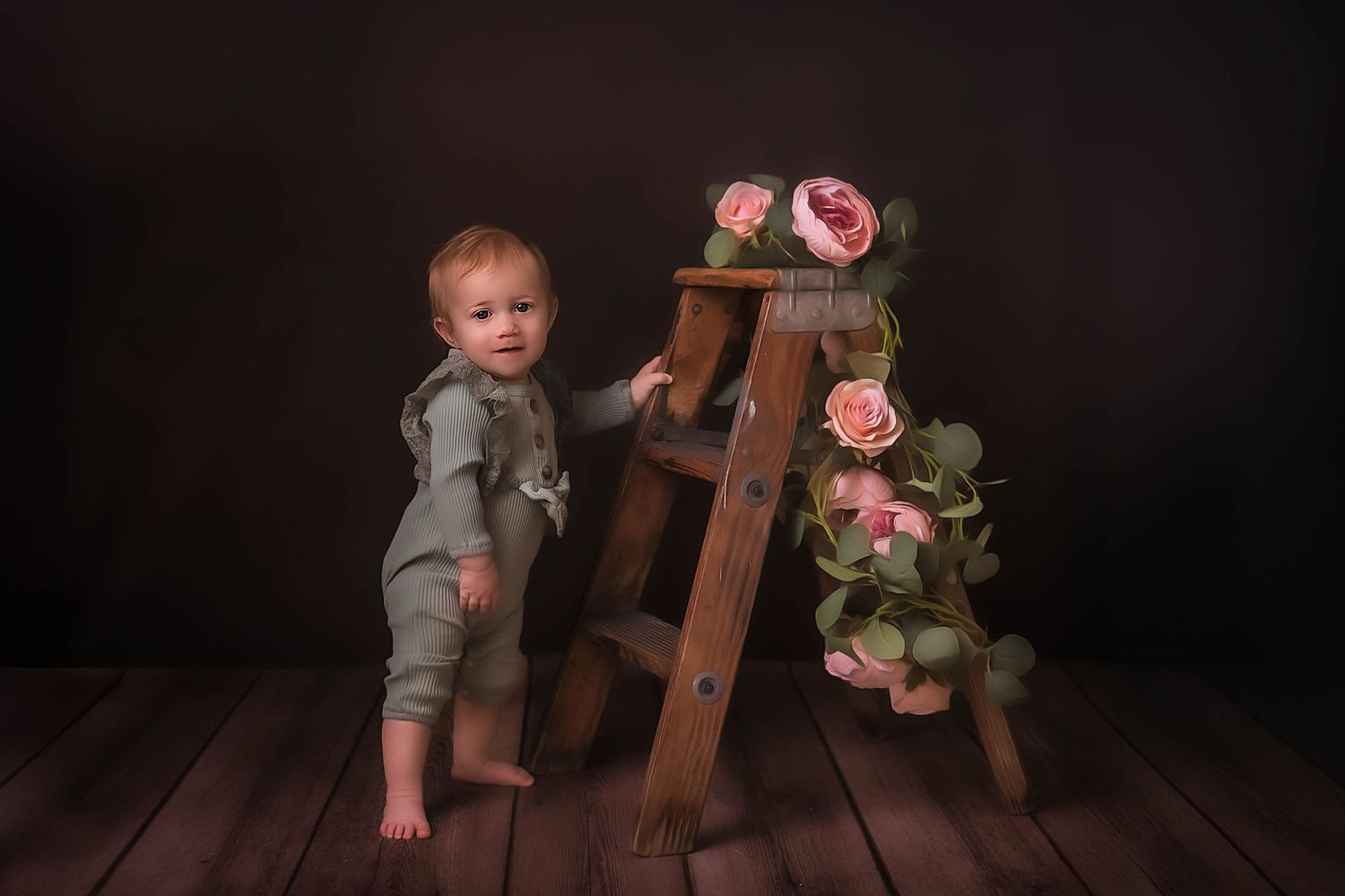 A small child in gray overalls stands beside a wooden ladder decorated with pink roses on a dark wooden floor in Kewaskum, WI Studio