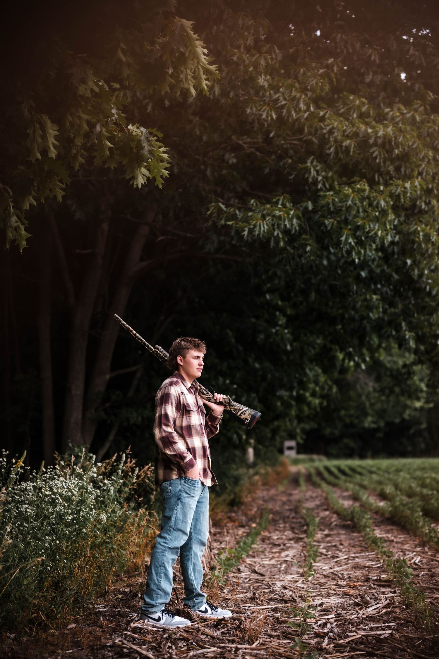 Young man in plaid shirt and jeans holding a rifle on a rural dirt path surrounded by green trees.