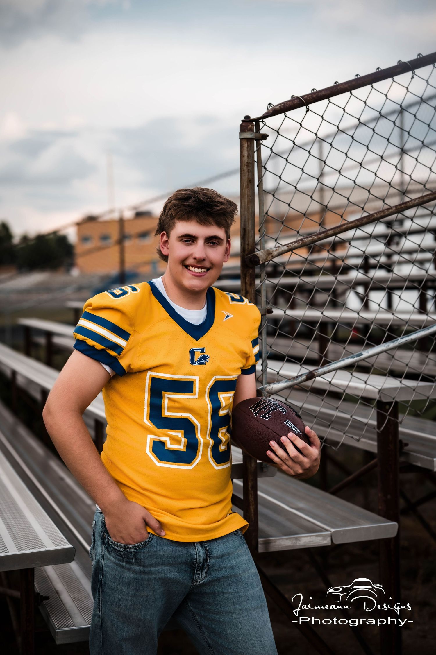 A smiling young man in a yellow football jersey number 58 holds a football while posing on bleachers.