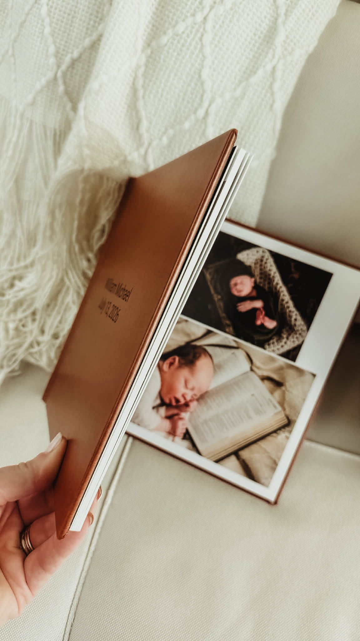 Hand holding an open leather-bound newborn photo album with engraved cover, showing sleeping baby photos beside a cozy cream knit blanket.