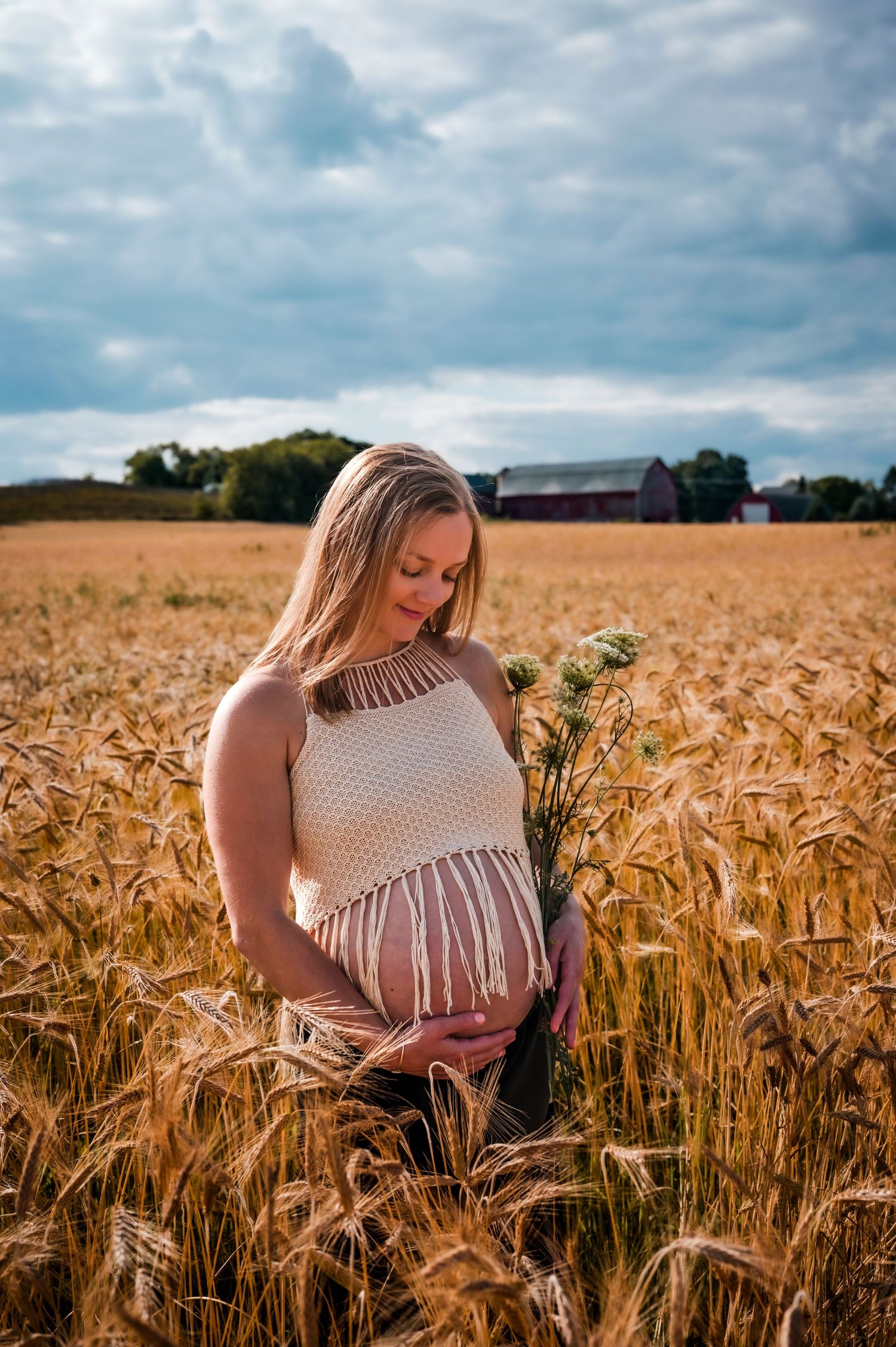 Pregnant woman in wheat field holding wildflowers, wearing crochet top, with red barn in background under cloudy sky.