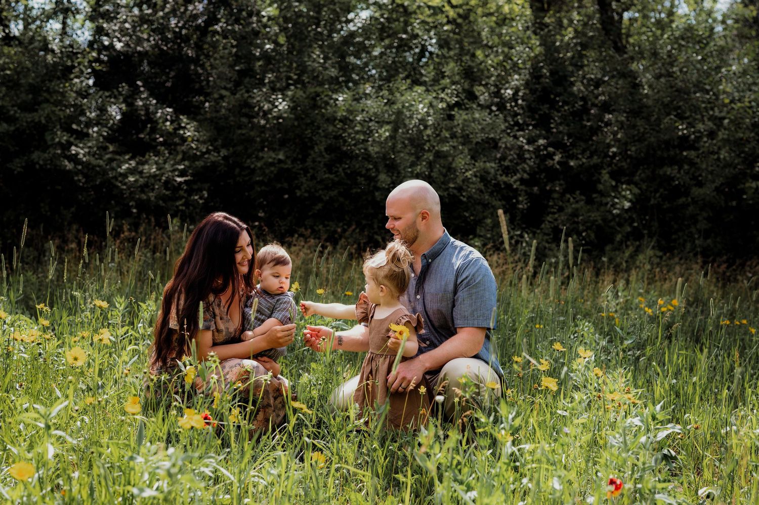 Family of four enjoying a sunny day in a wildflower meadow, with two young children and lush green trees in the background.