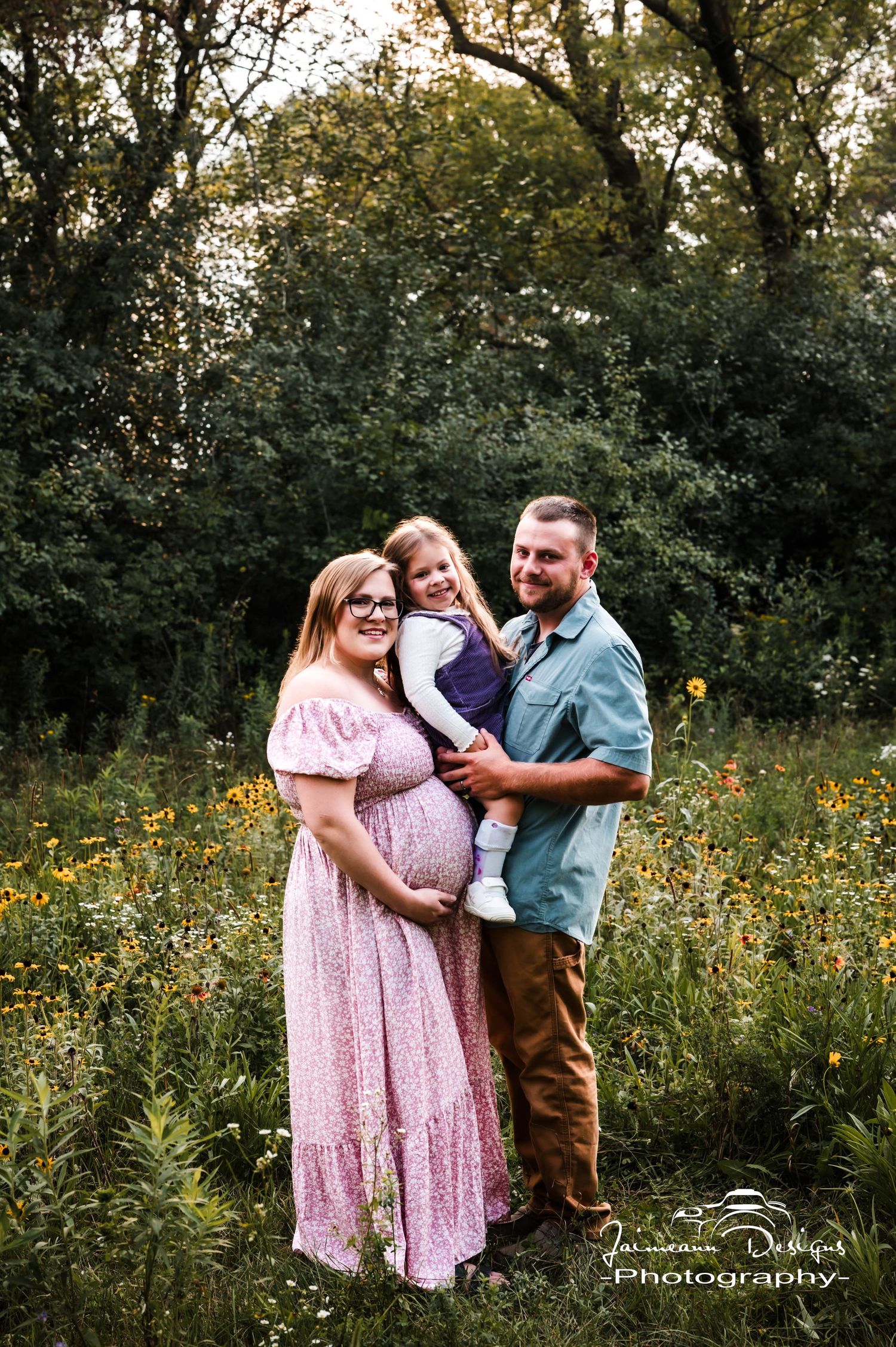 Pregnant woman in pink dress poses with husband and young child in a wildflower meadow for a maternity photo session.