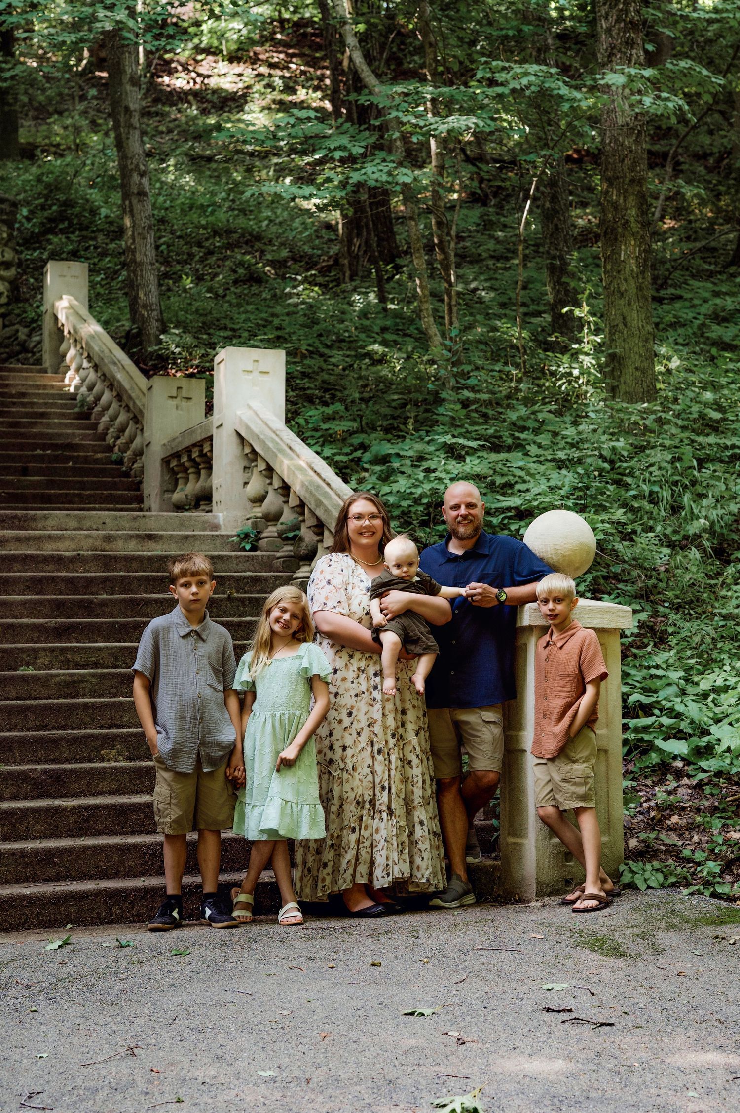 Family of six posing on stone staircase path surrounded by lush green forest trees during summer outdoor photo session.
