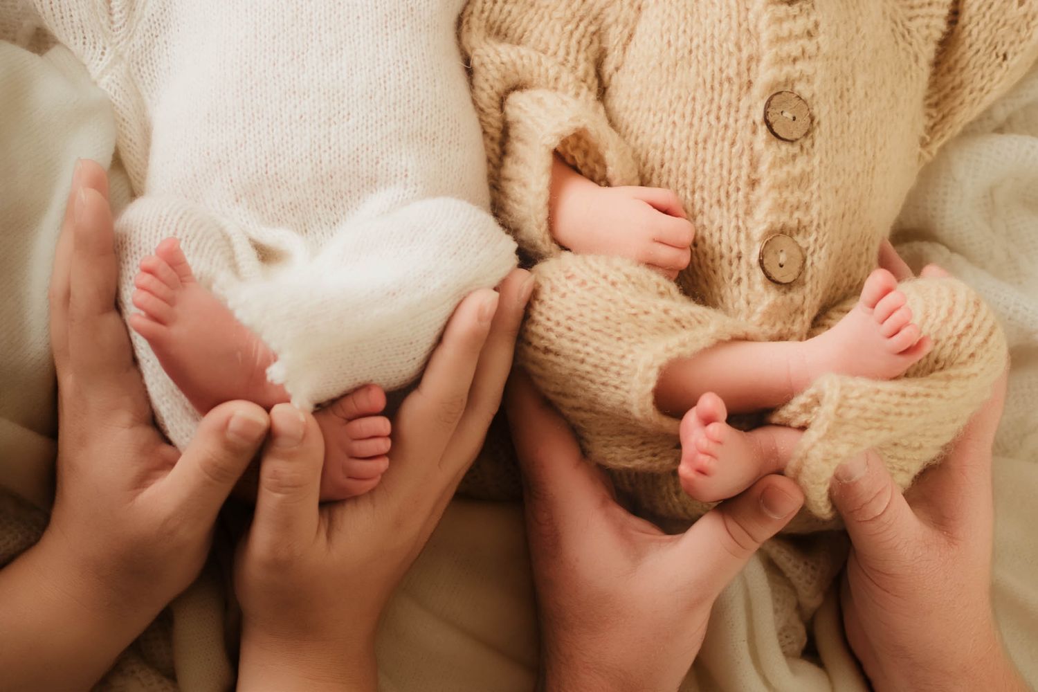 Two newborn babies' tiny feet resting in parents' cupped hands, one in white swaddle and one in a cozy knit cardigan.