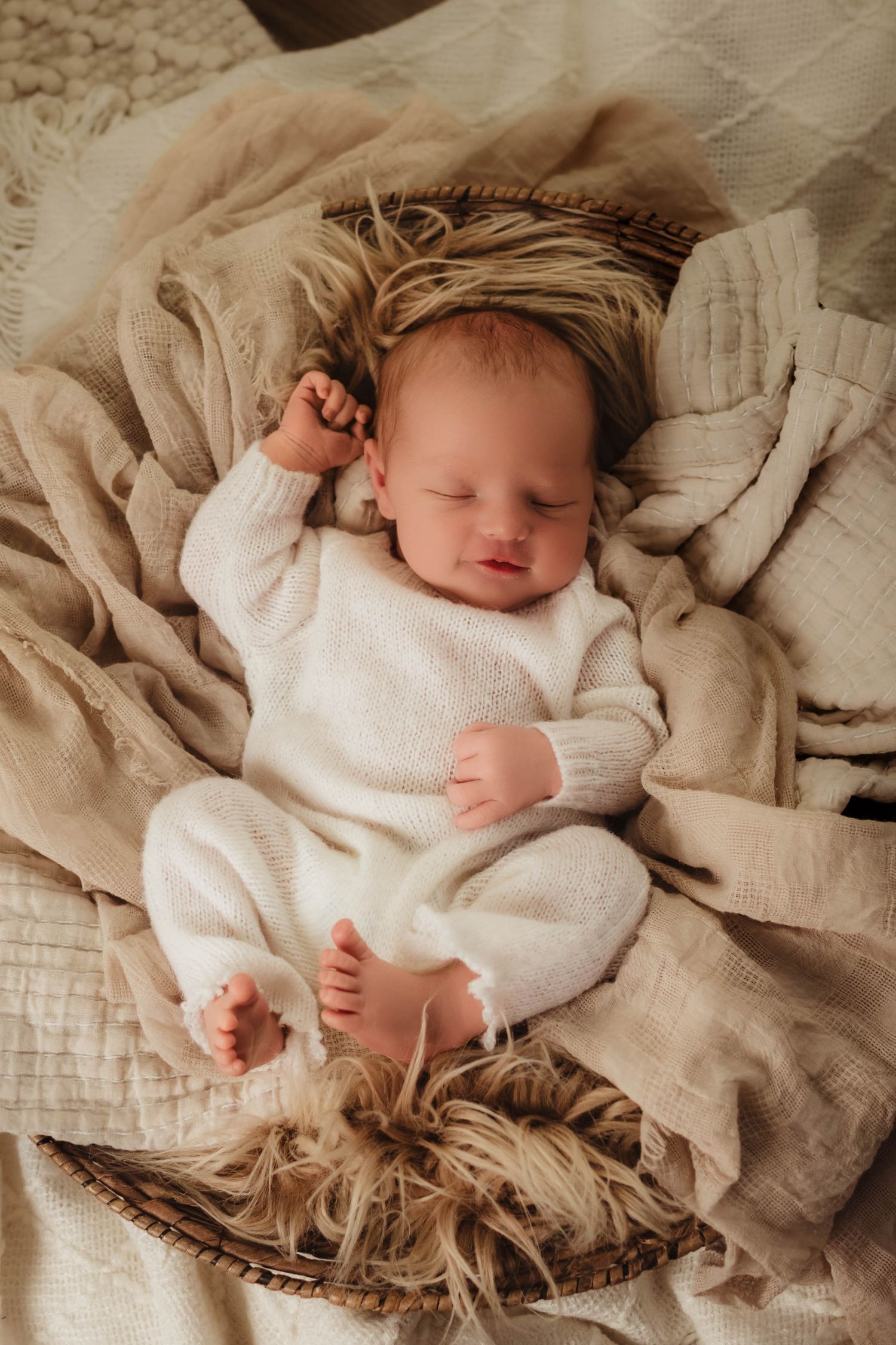 Sleeping newborn baby in a cream knit outfit lying on a woven basket with soft neutral blankets and fluffy faux fur.