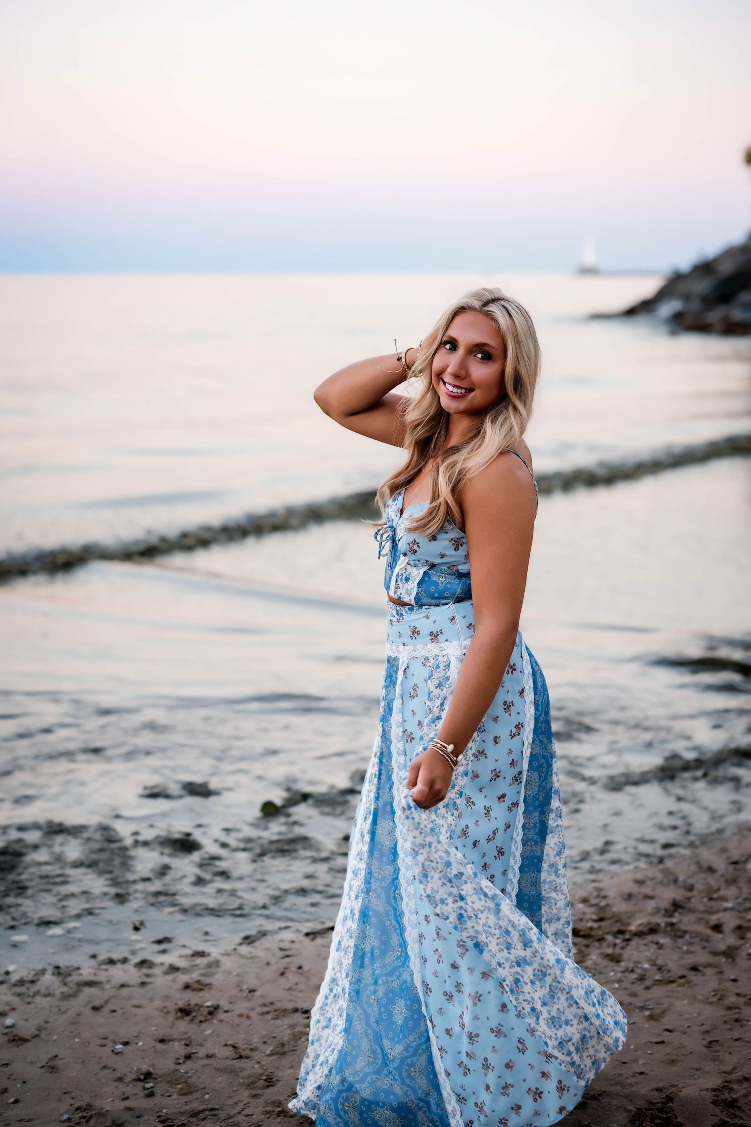 Senior girl holding her blue floral maxi dress on a Lake Michigan beach at sunset