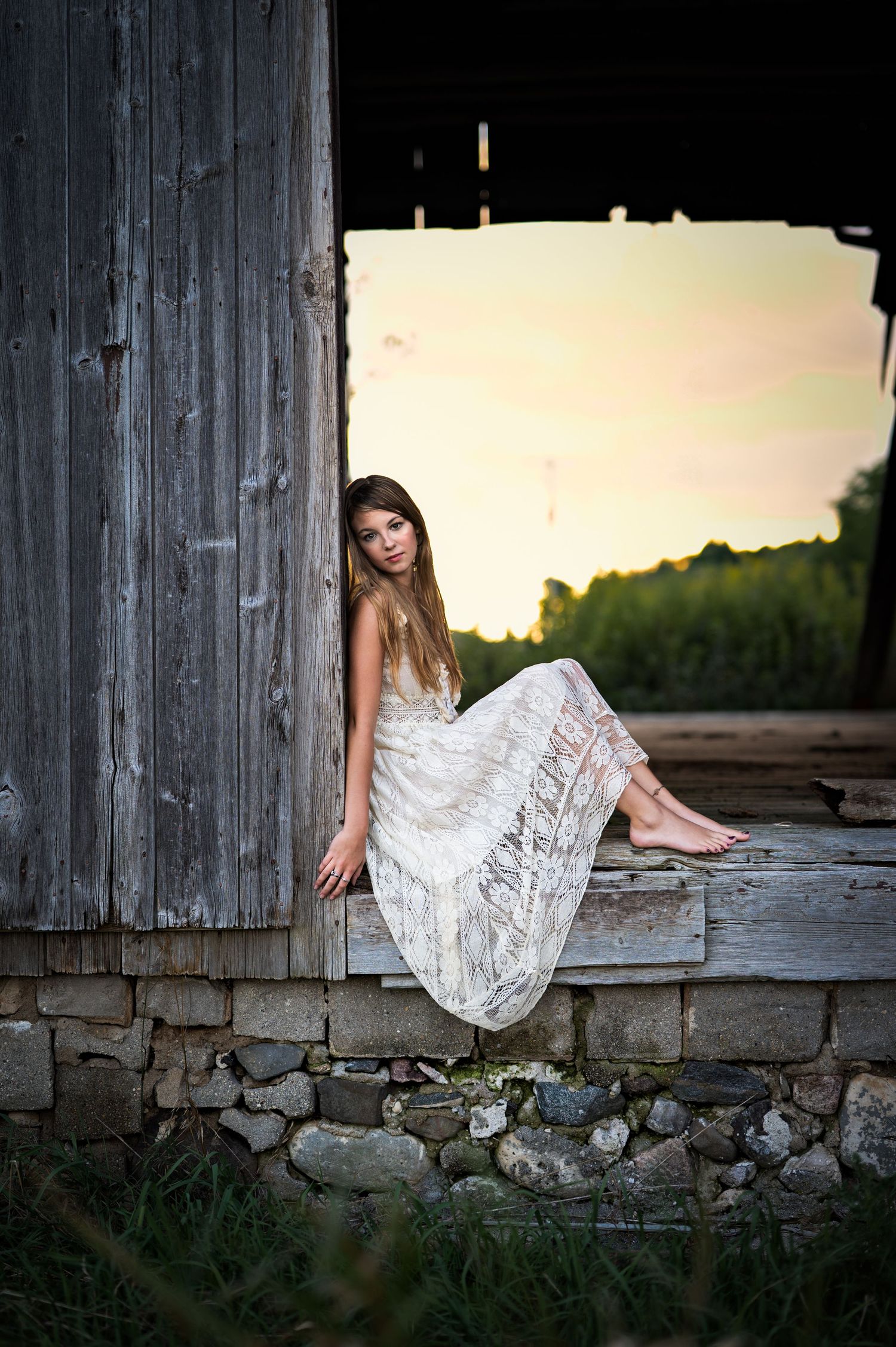 A person in a white lace dress poses gracefully on a rustic stone wall at sunset in a series of artistic photos in Plymouth, WI