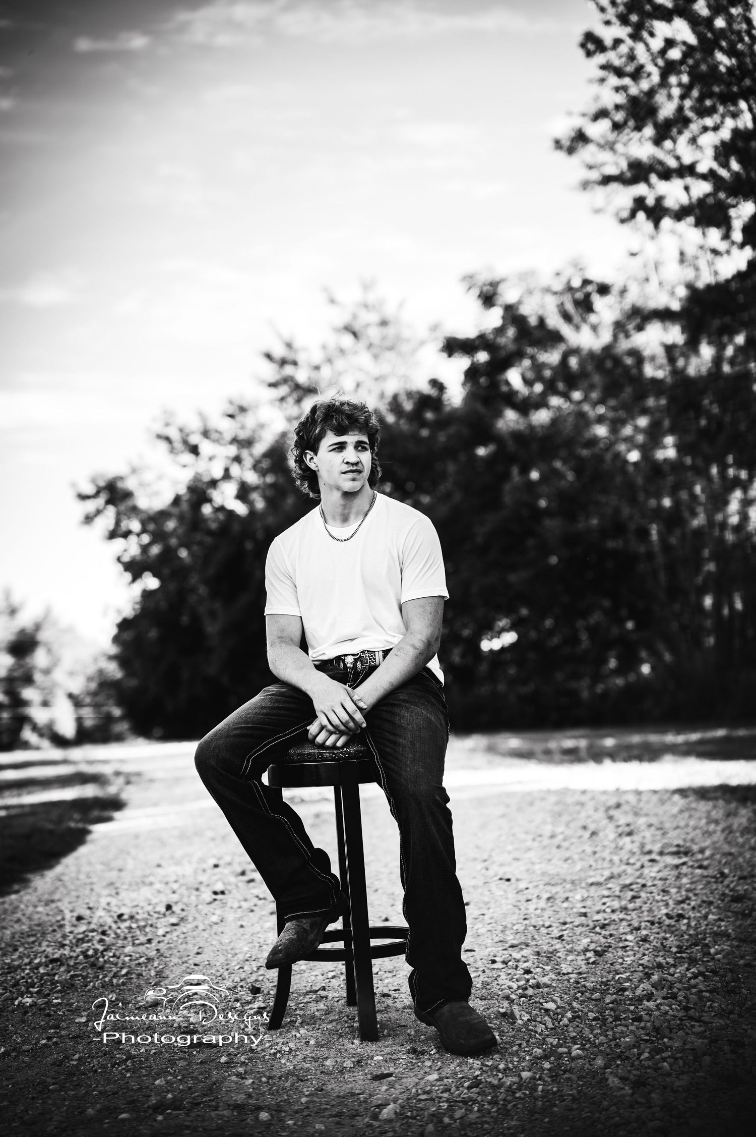 Black and white portrait of someone sitting on stool outdoors in casual attire.