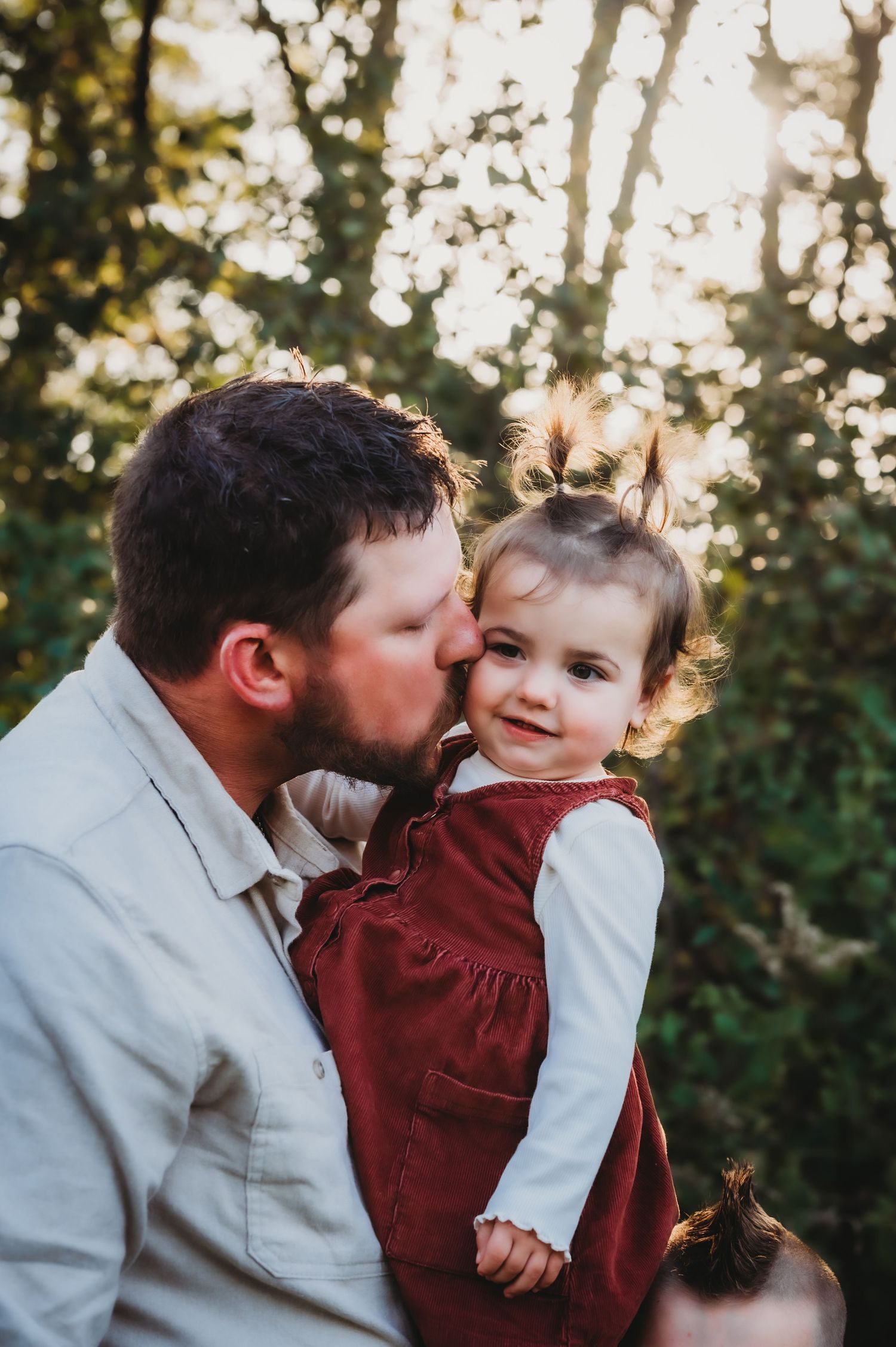 Outdoor portrait of gentle moment between parent and toddler in burgundy dress against sunlit foliage background.