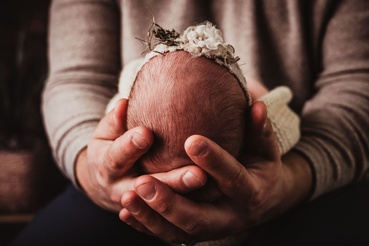 Gentle hands cradling newborn's head adorned with delicate floral headband.
