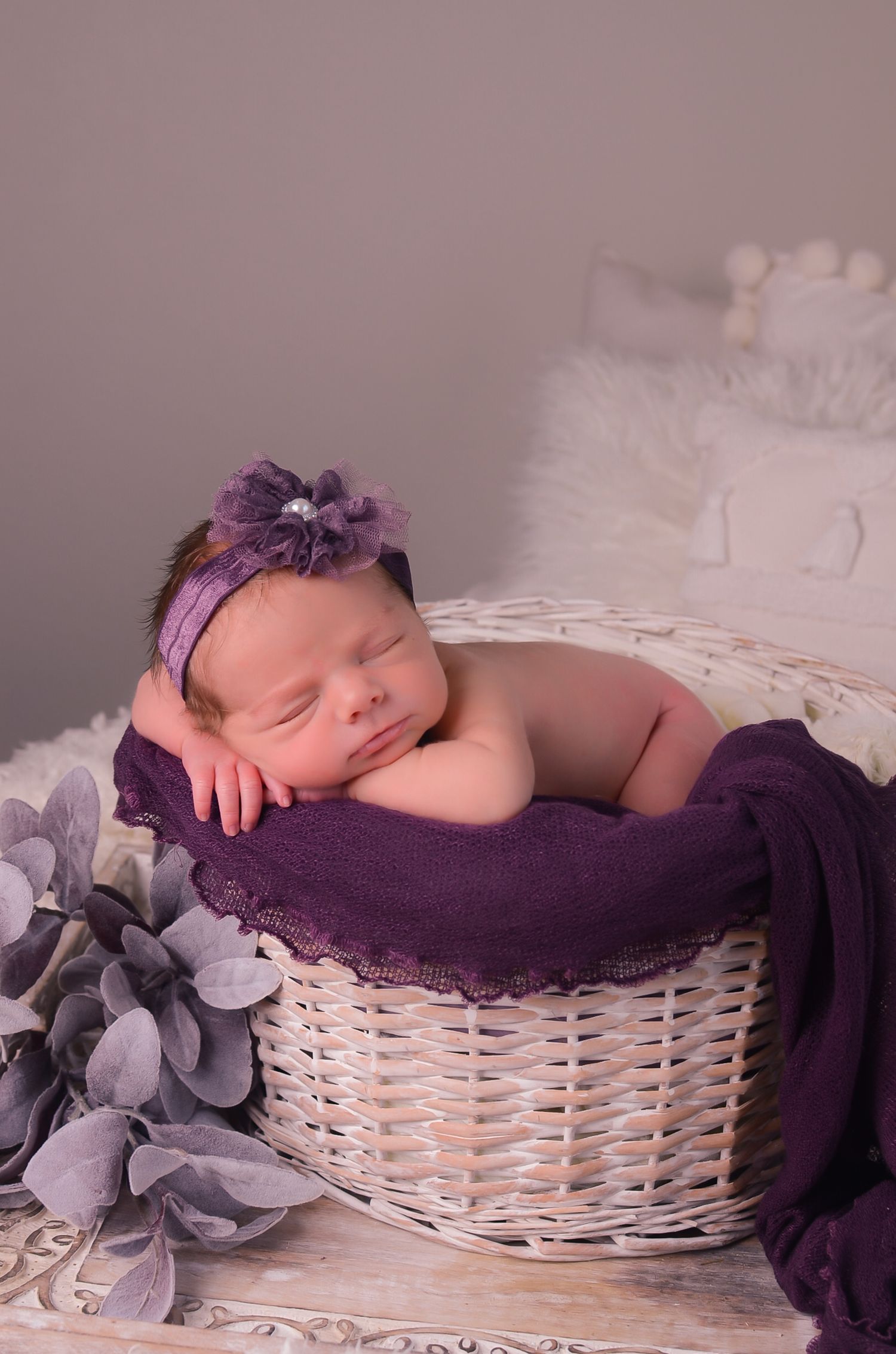 A sleeping baby wrapped in purple fabric rests peacefully in a white woven basket with soft floral decorations.