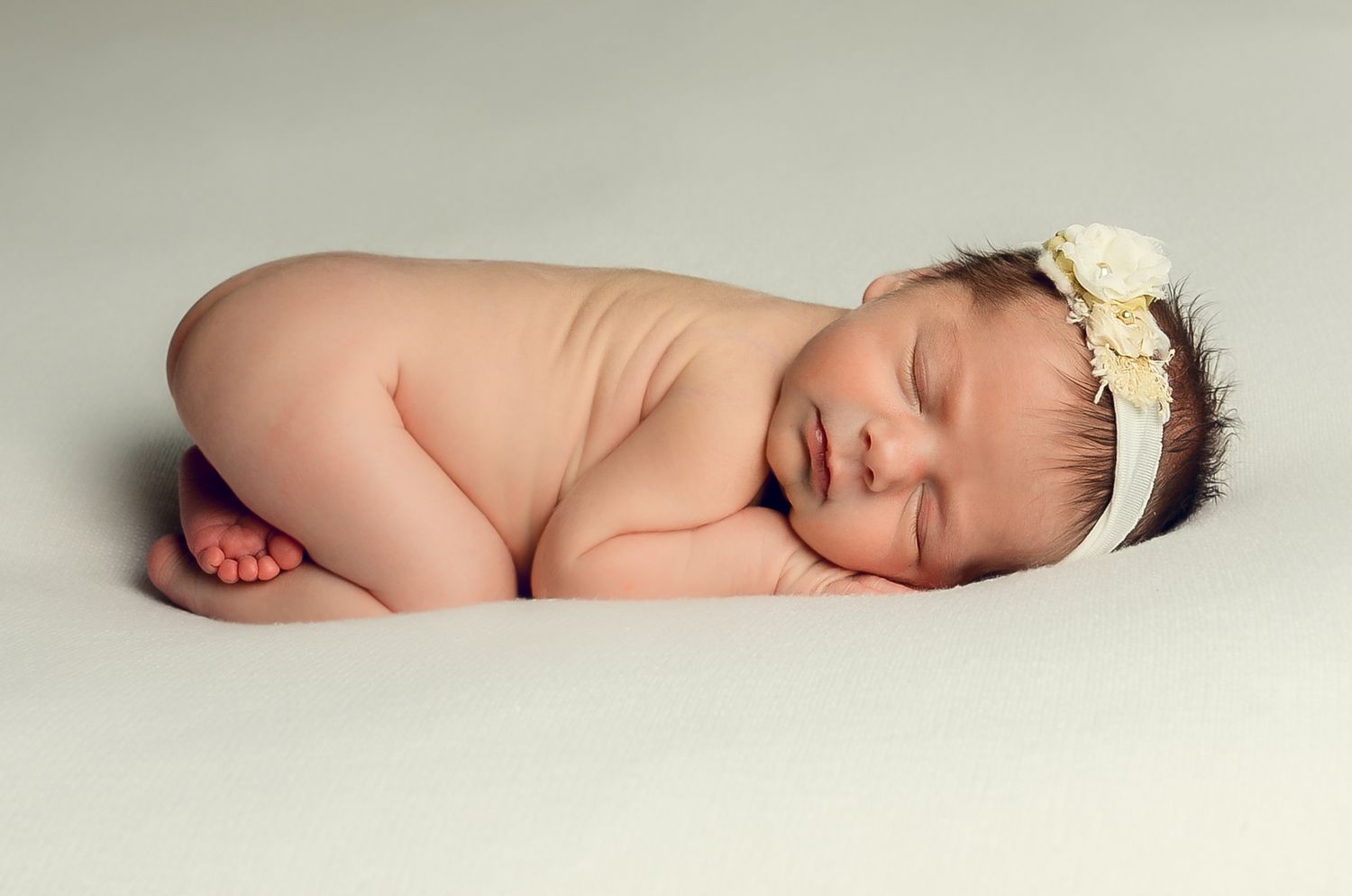 Peaceful sleeping newborn baby wearing a delicate white floral headband on cream backdrop. Cedarburg, WI