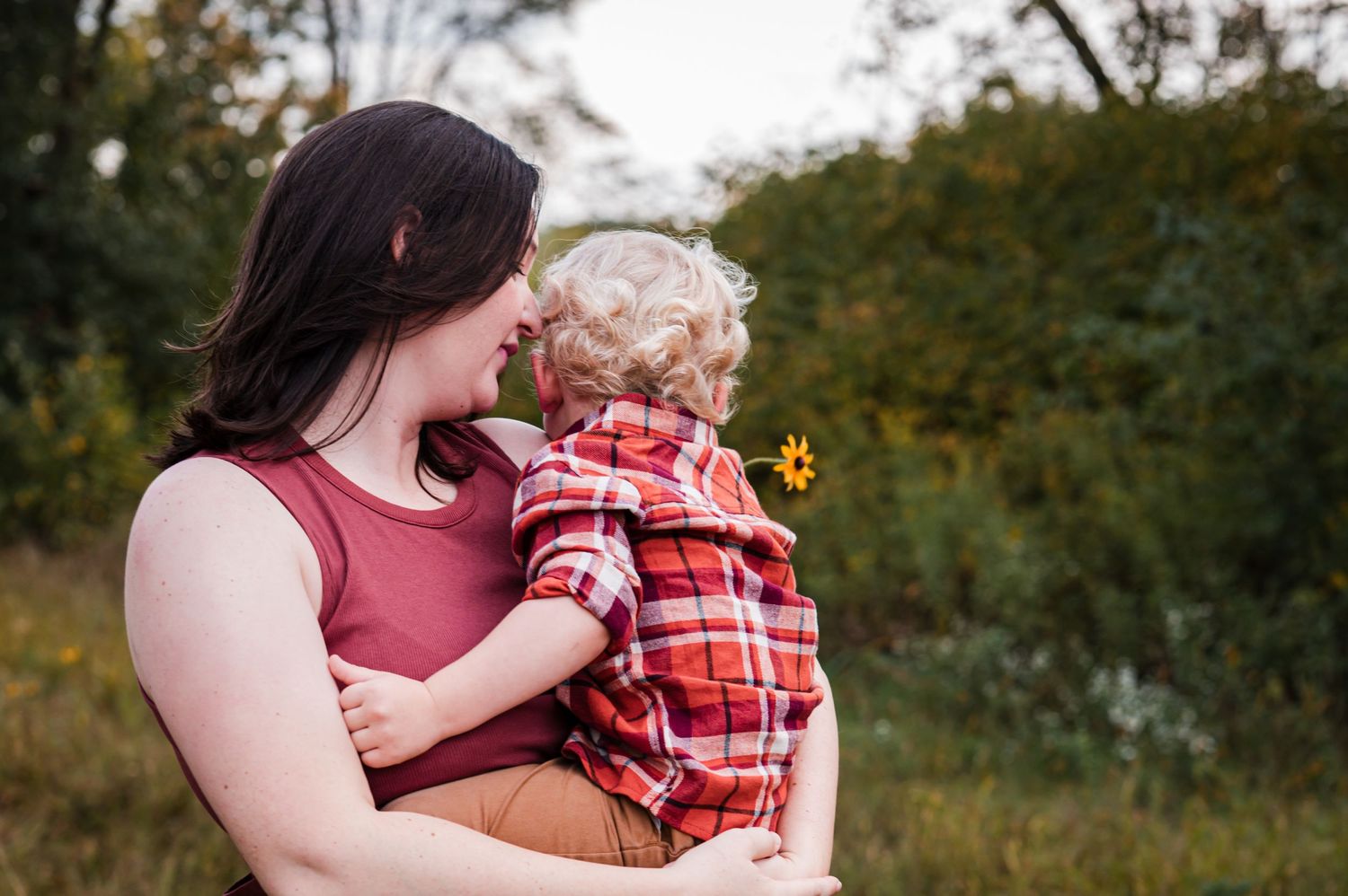 A mother in a burgundy top holds and nurtures her baby wearing a red plaid outfit outdoors. Campbellsport, WI location