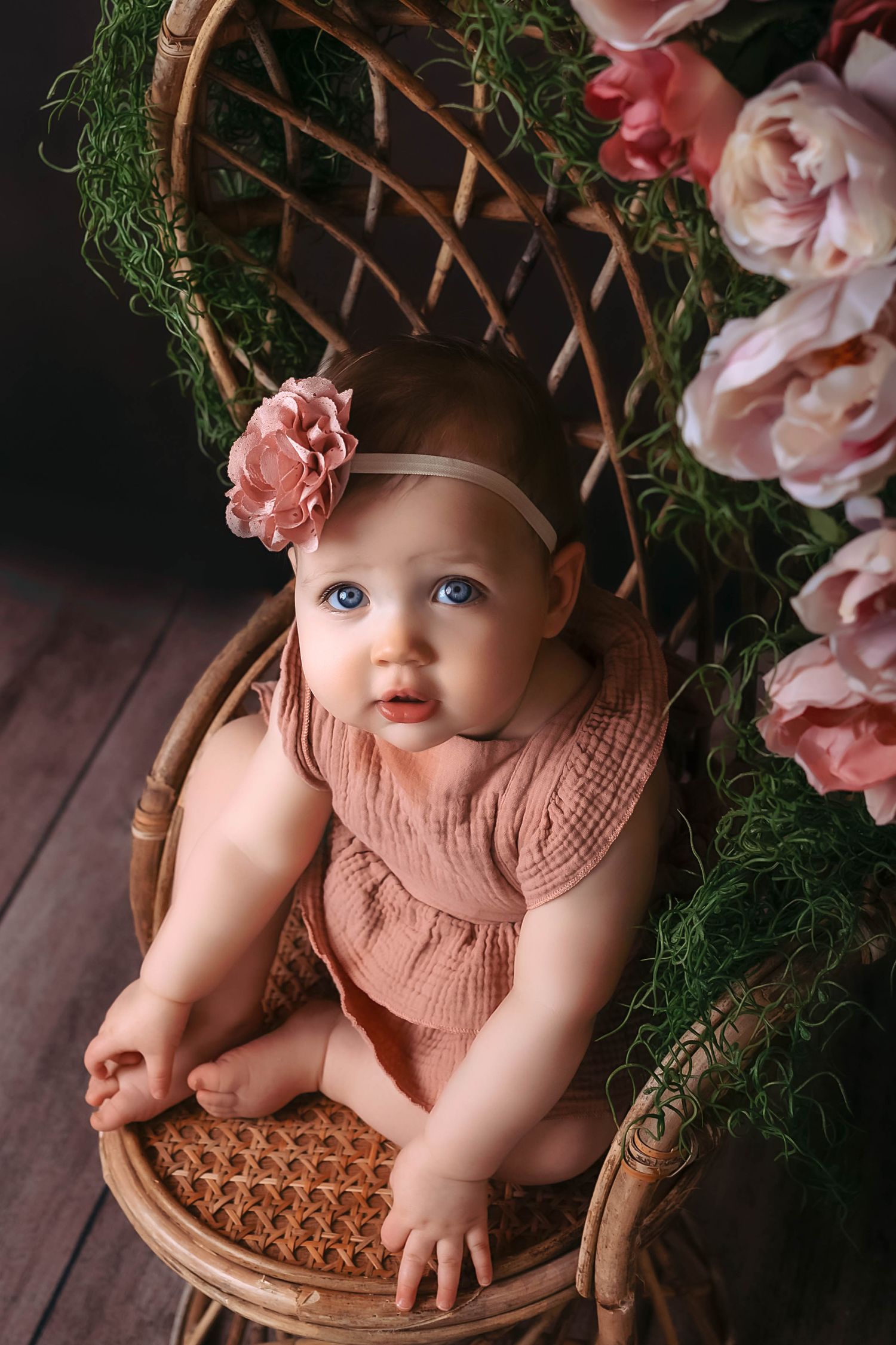 Baby in pink dress sits in wooden basket surrounded by floral decorations and greenery. One Year Milestone Session, Kewaskum, WI Studio