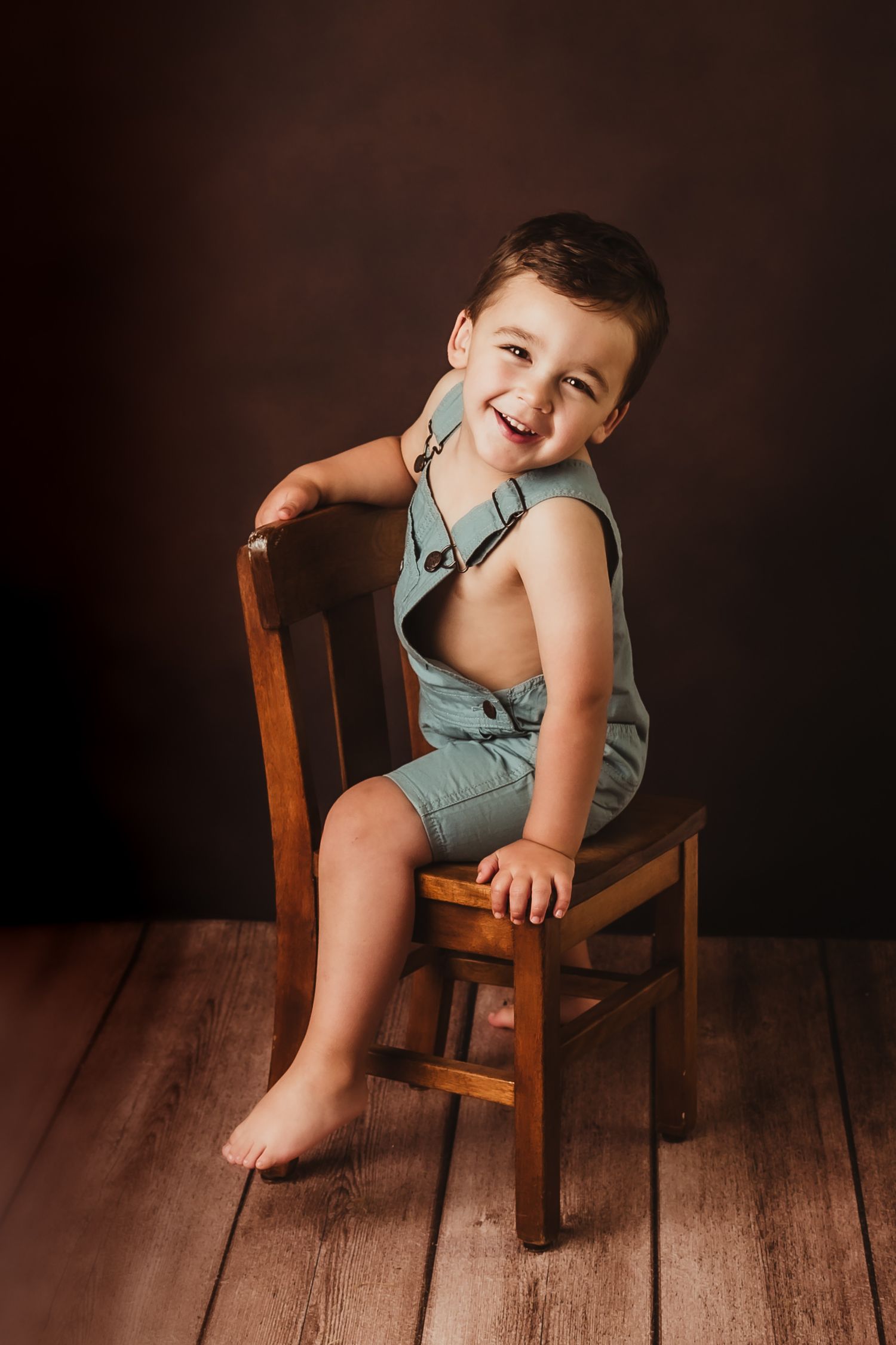 Joyful child wearing mint green overalls sits on wooden chair against dark backdrop in studio milestone portrait, WI