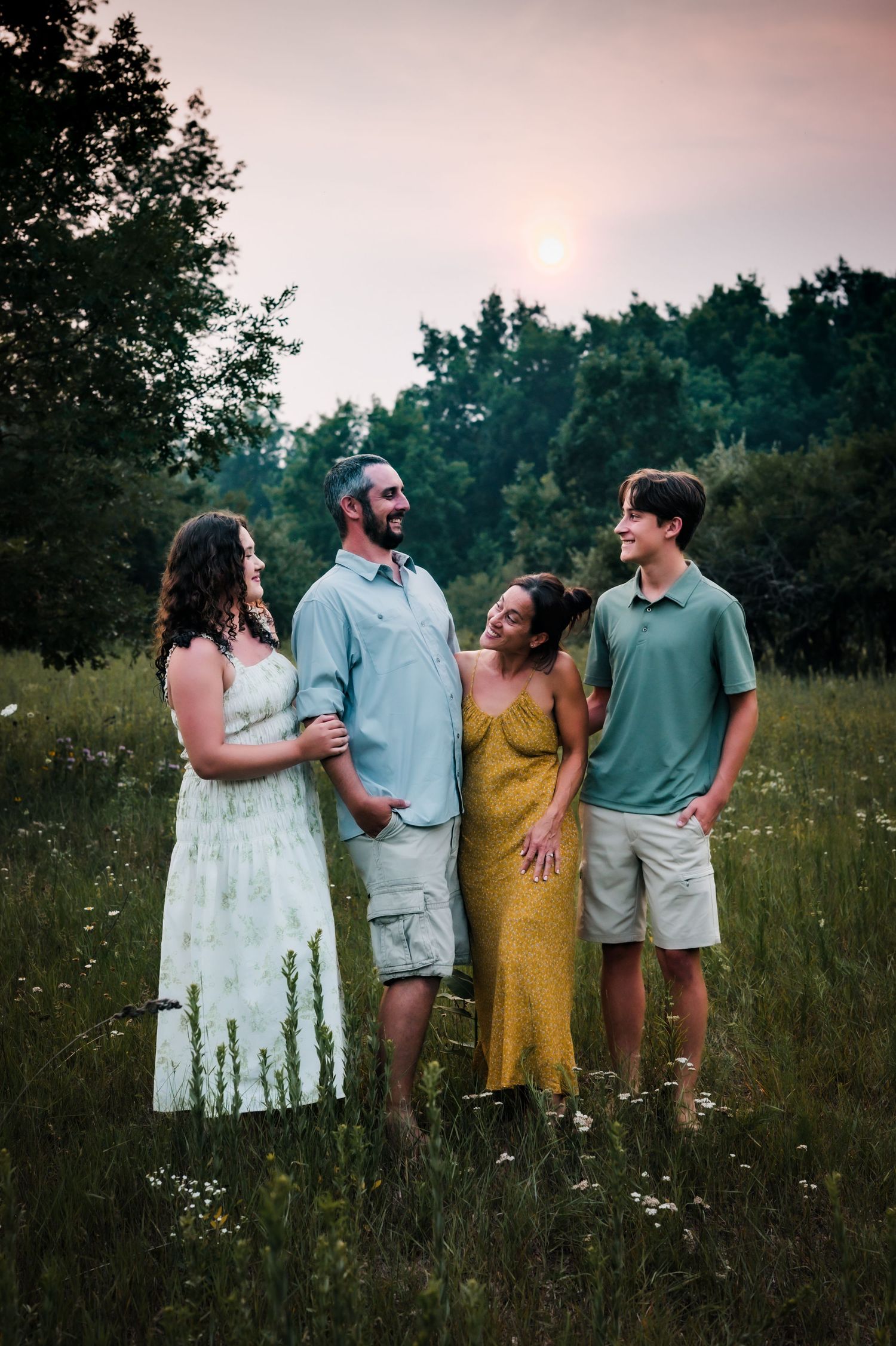 Family of four smiling together in a wildflower meadow at sunset, wearing casual summer outfits in soft greens and yellow.