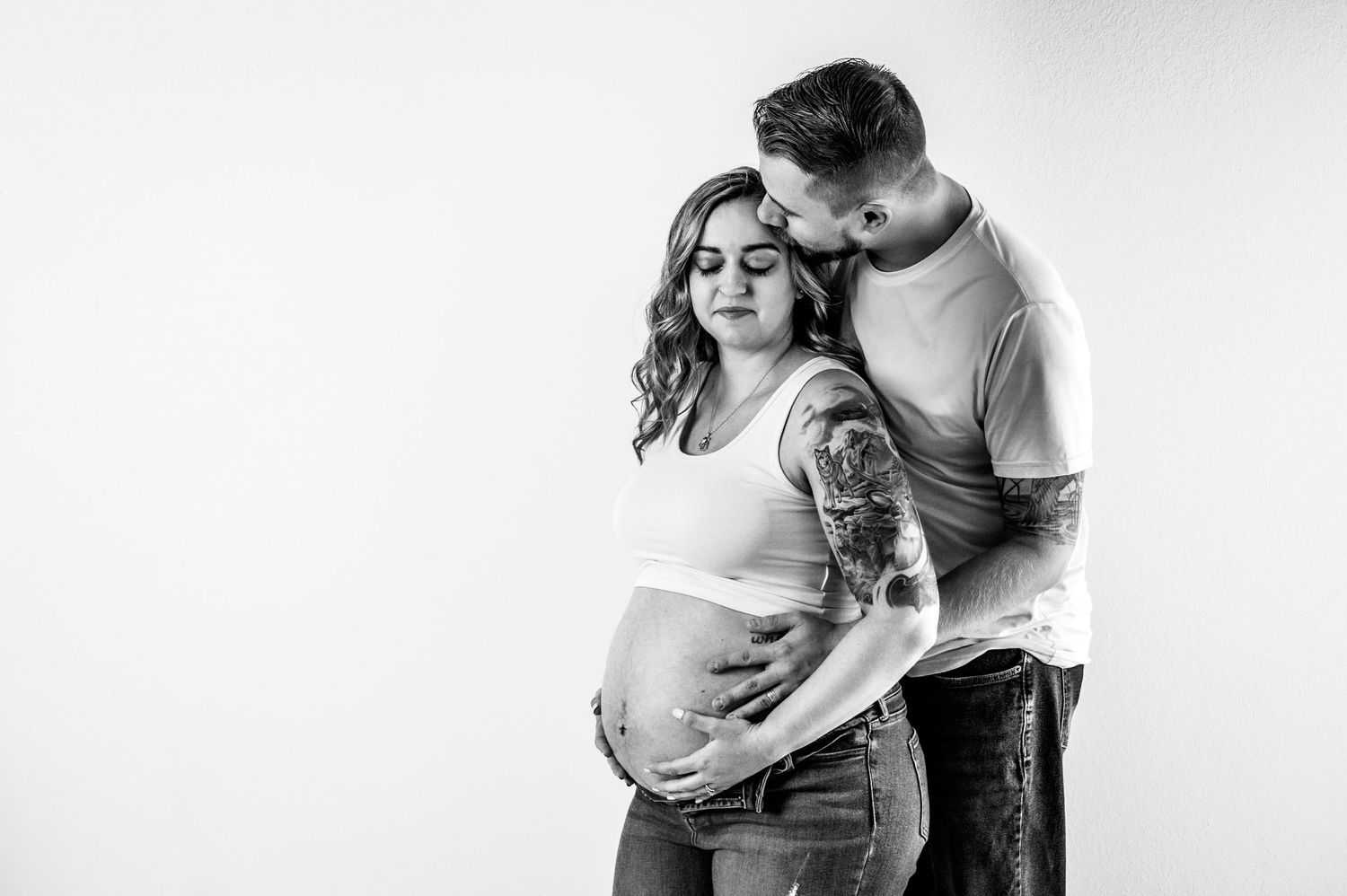 Black and white maternity photo of pregnant woman and tattooed partner embracing, both looking down at baby bump.