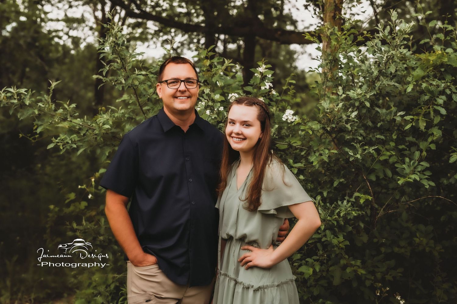 Couple poses outdoors for portrait in casual attire against green foliage background.