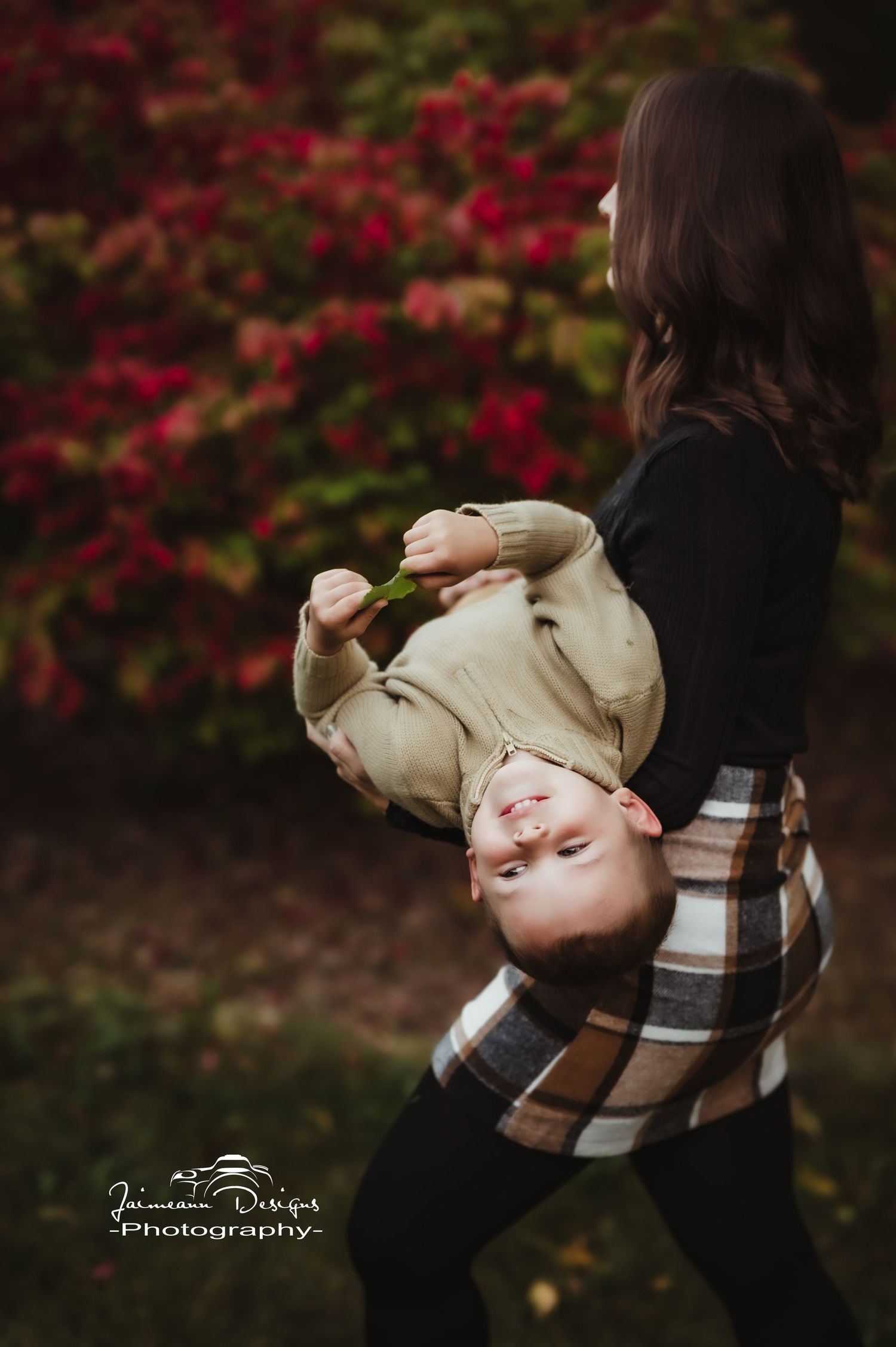 Playful moment captured as parent swings child upside down against red autumn bushes.