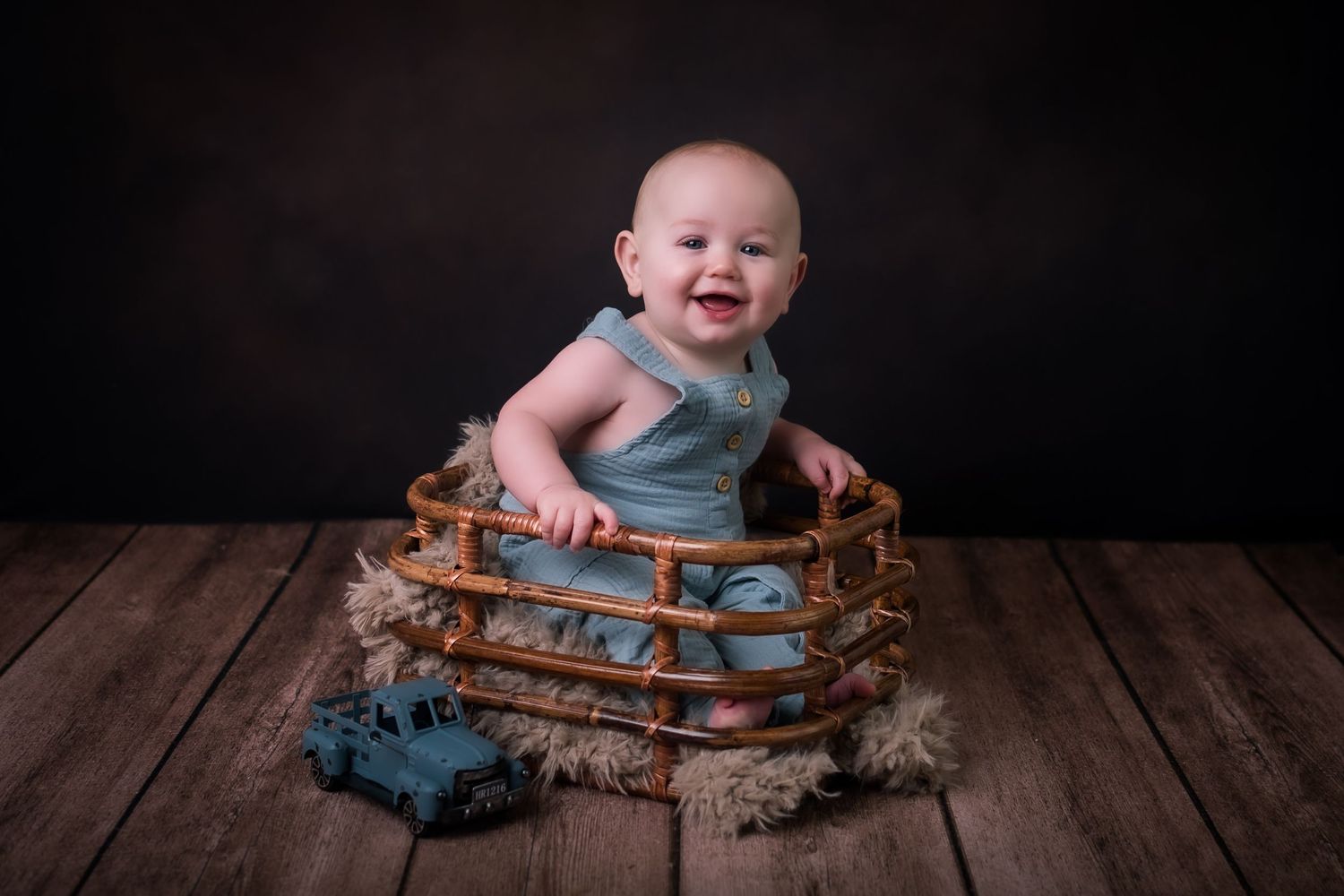 A smiling baby sits in wooden crate surrounded by toy dinosaurs against dark background.