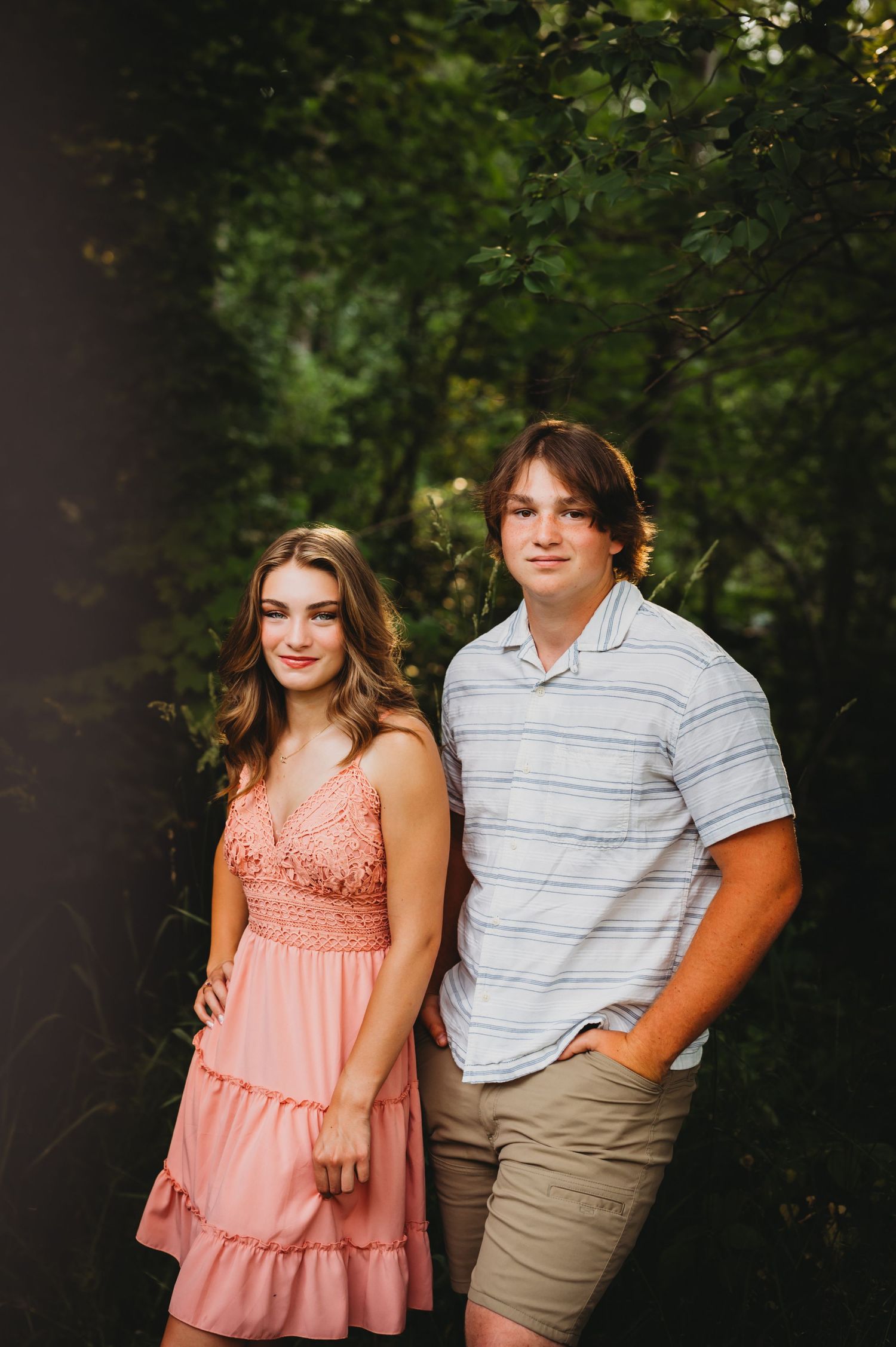 Couple poses together outdoors, wearing pink dress and striped shirt against green foliage.
