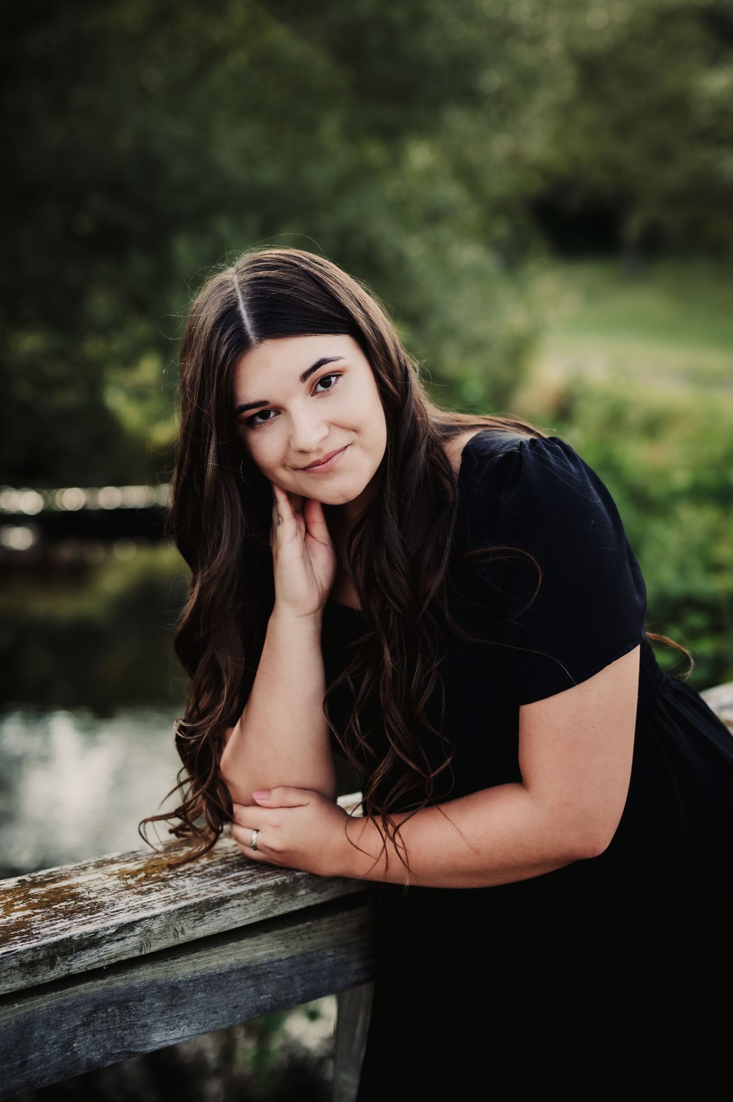 Dark-haired figure in black dress poses by a wooden railing near a pond.