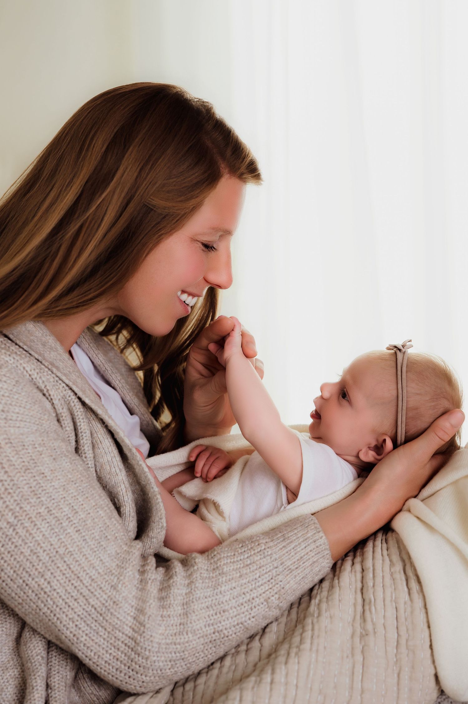 Smiling young mother holding her newborn baby girl in a white blanket, sharing a tender moment together.