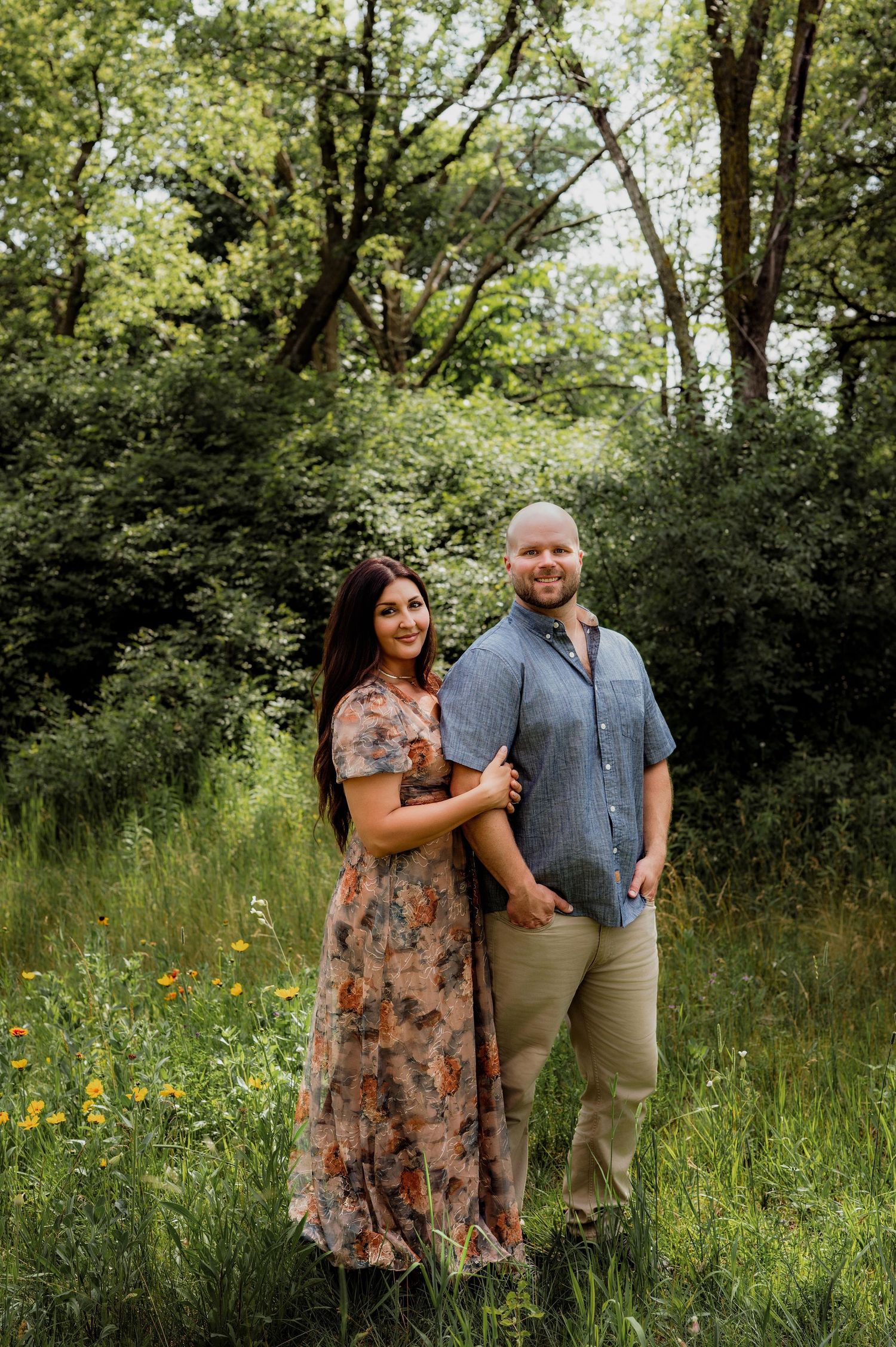 Couple posing in a lush green meadow, woman in floral maxi dress and man in blue shirt, surrounded by trees.