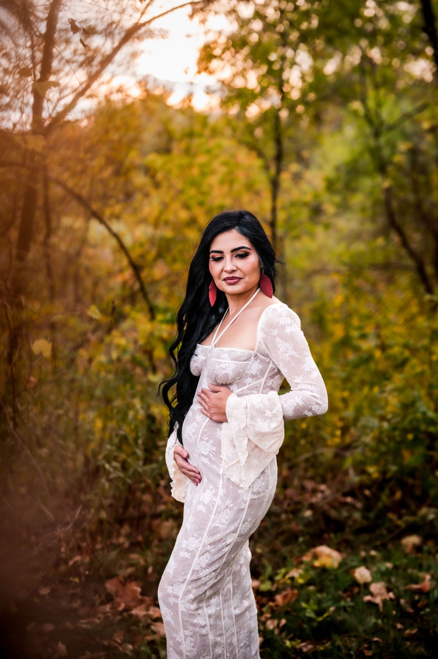 Pregnant woman in white lace gown with red earrings poses in autumn forest during golden hour maternity photoshoot.