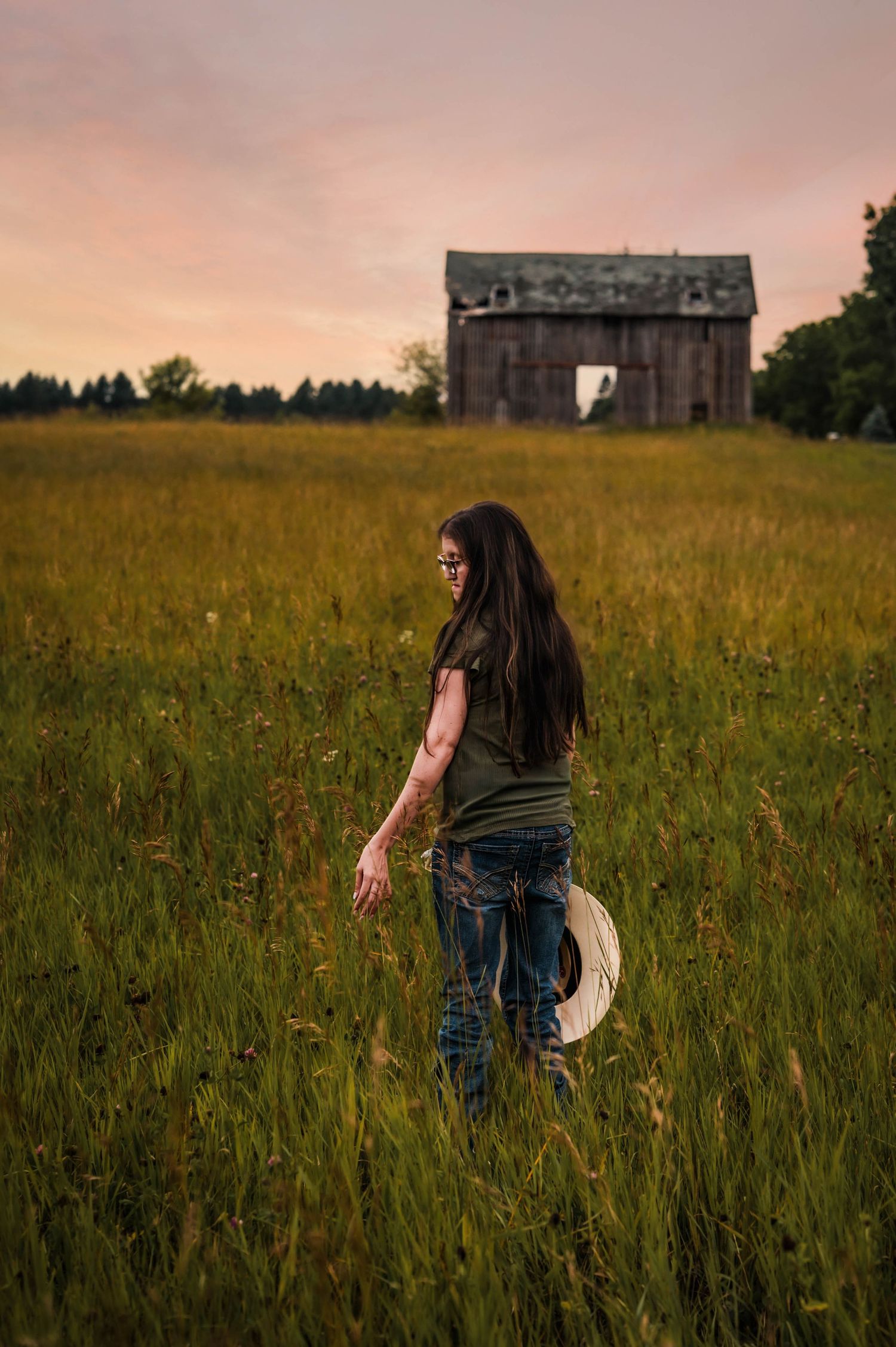 Woman with long hair holding a hat walks through tall grass at sunset with a rustic barn in the background.