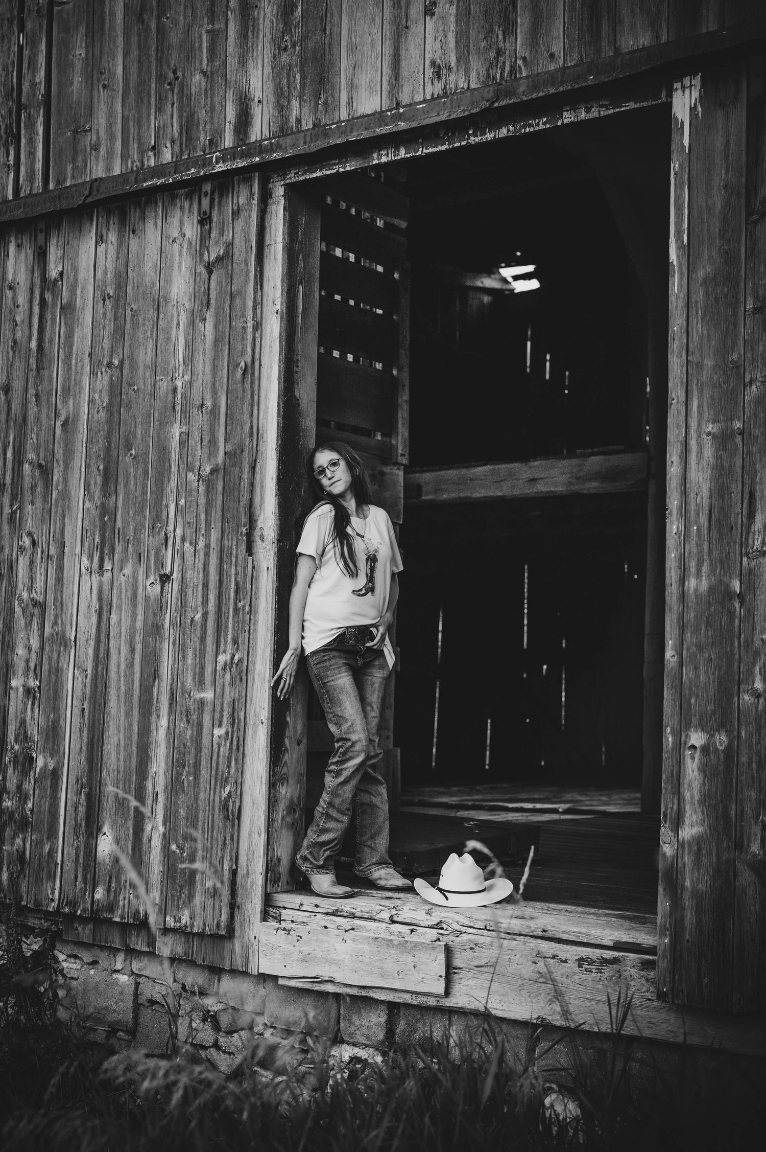 Black and white photo of a young woman leaning in an old wooden barn doorway with a cowboy hat at her feet.