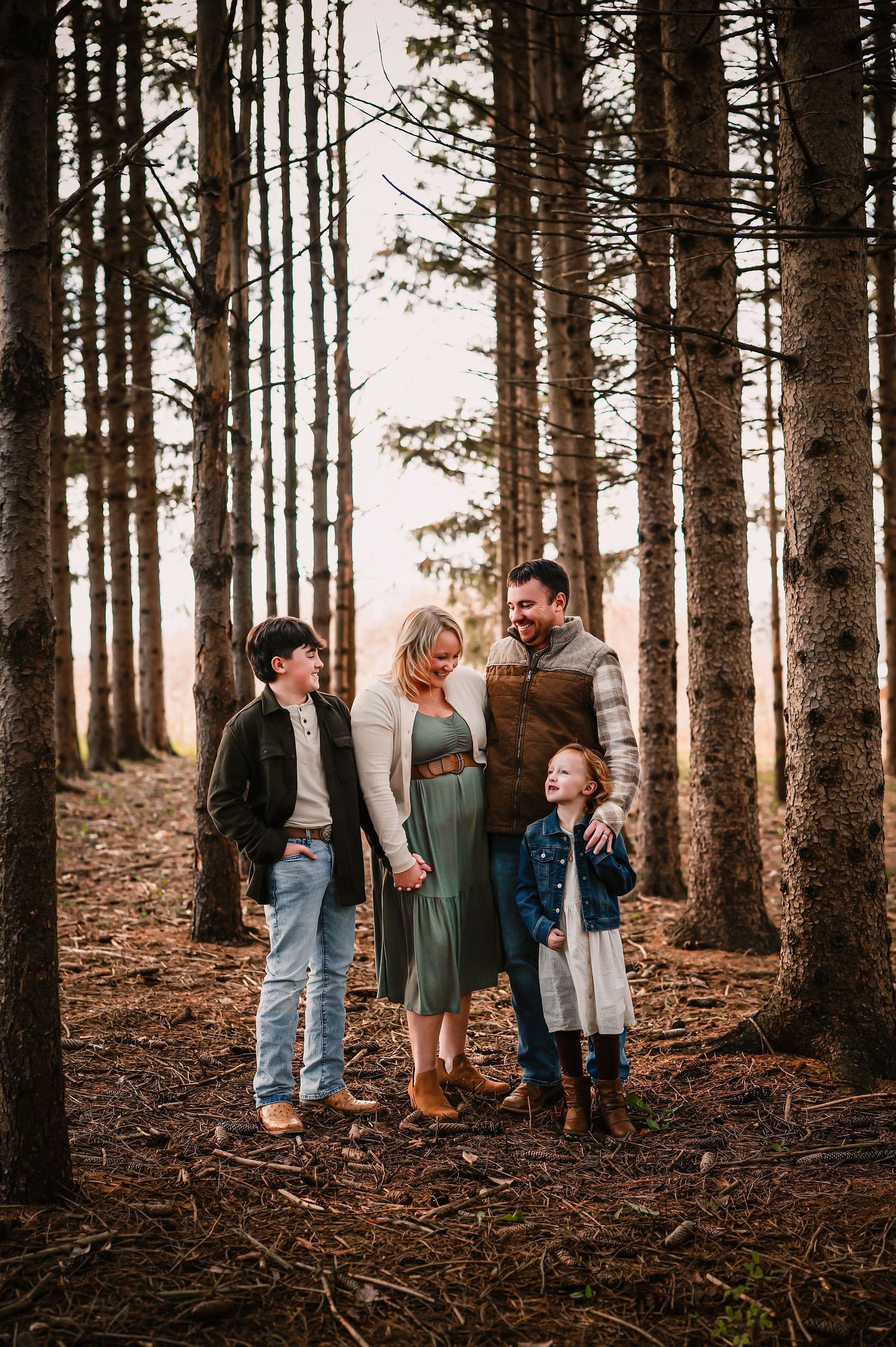 Family of four smiling together among tall pine trees in a forest during golden hour.