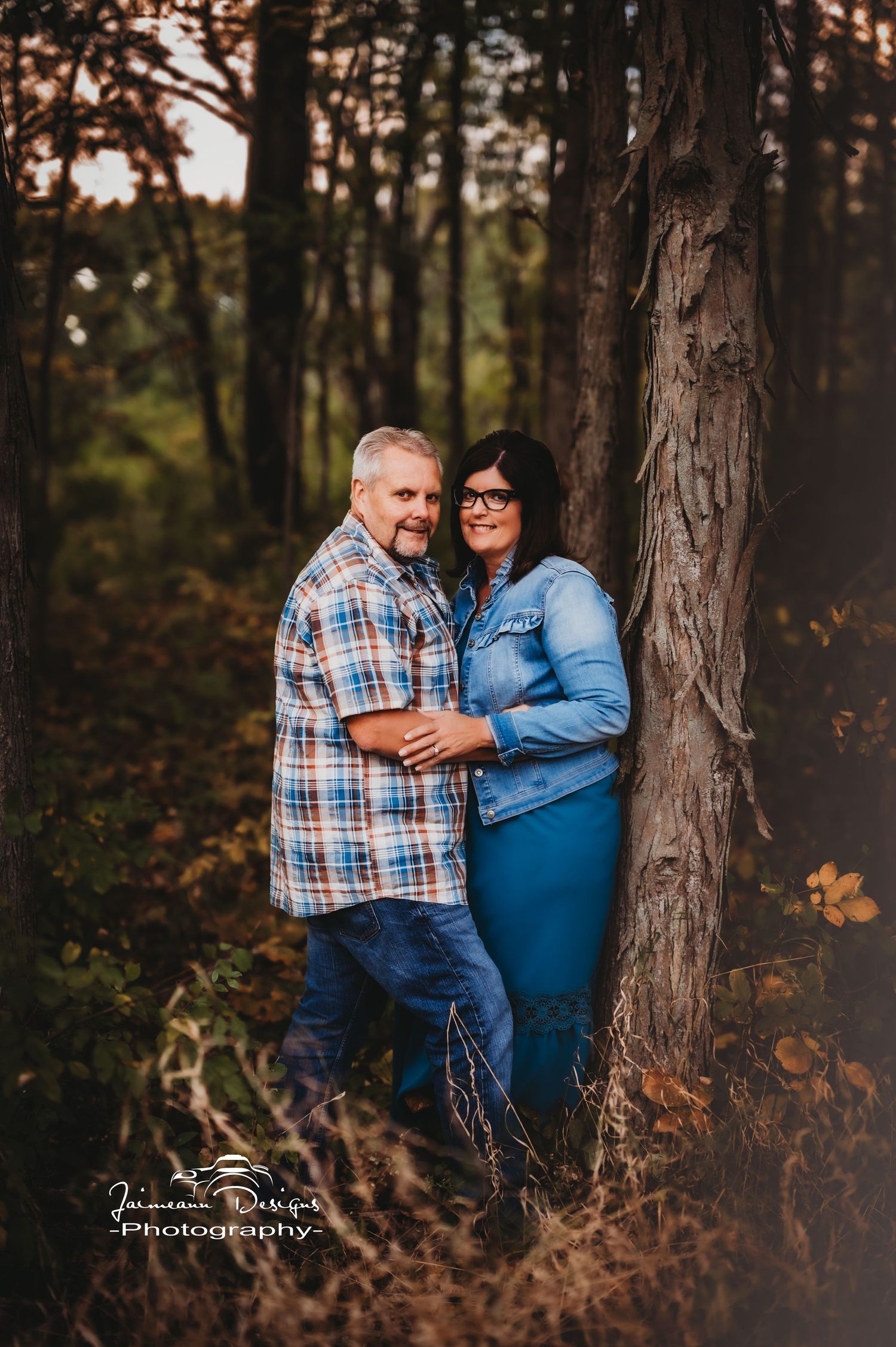 Couple posing intimately against large tree trunk in wooded forest setting.