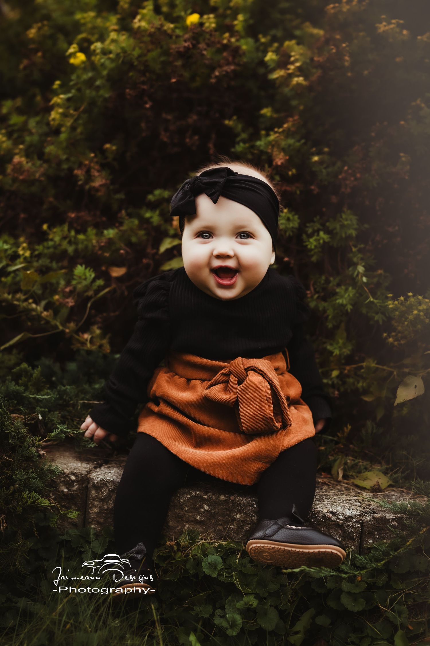 Baby in black and orange outfit sits on stone step with adorable expression.