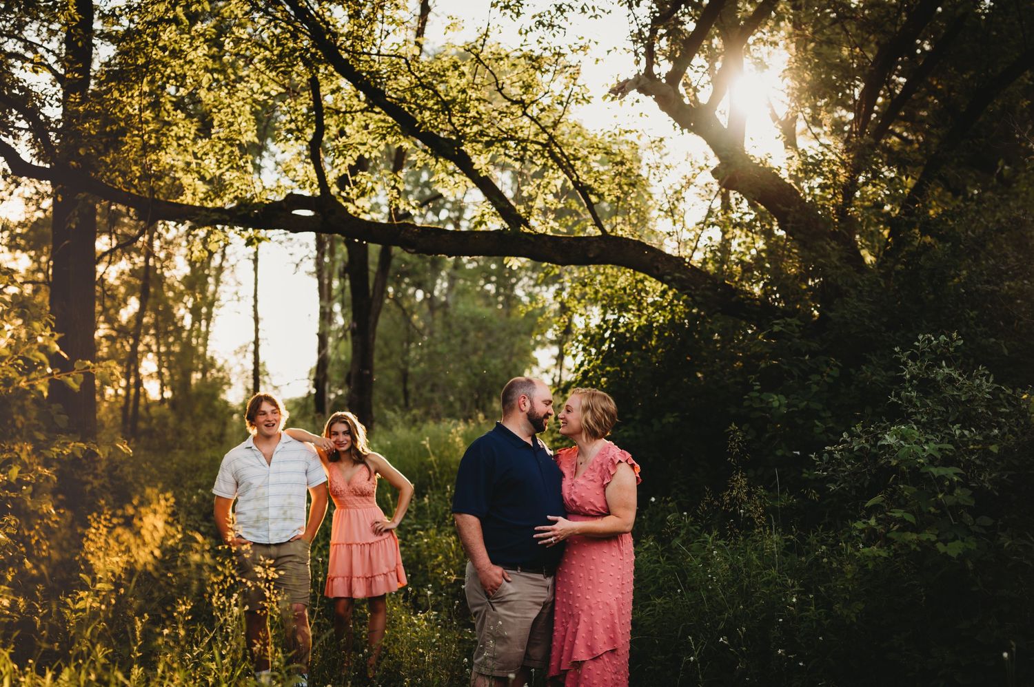 Two couples pose in wooded setting with golden sunlight filtering through trees.