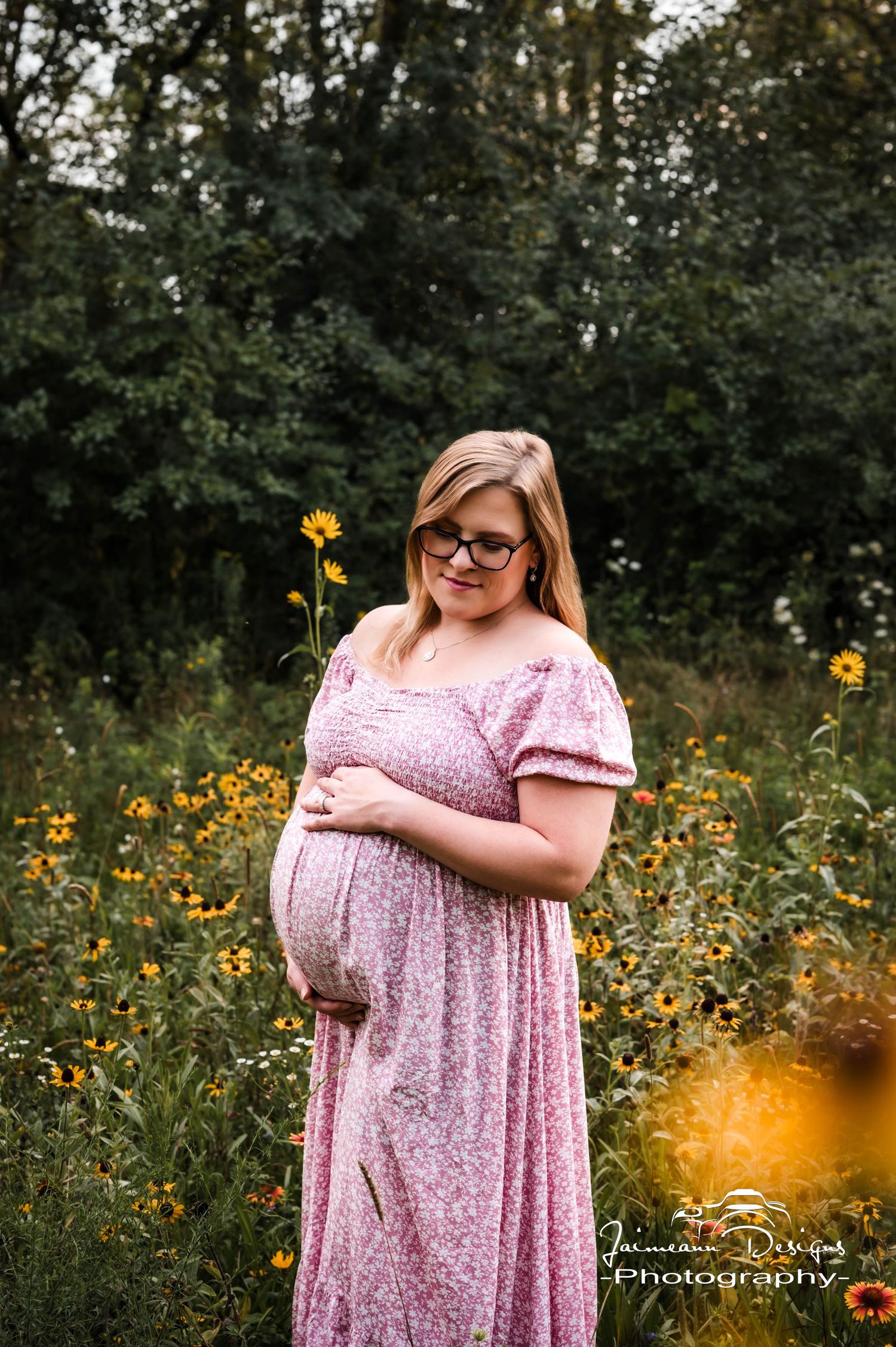Pregnant woman in pink floral dress holding belly in sunflower field, maternity photography outdoors.