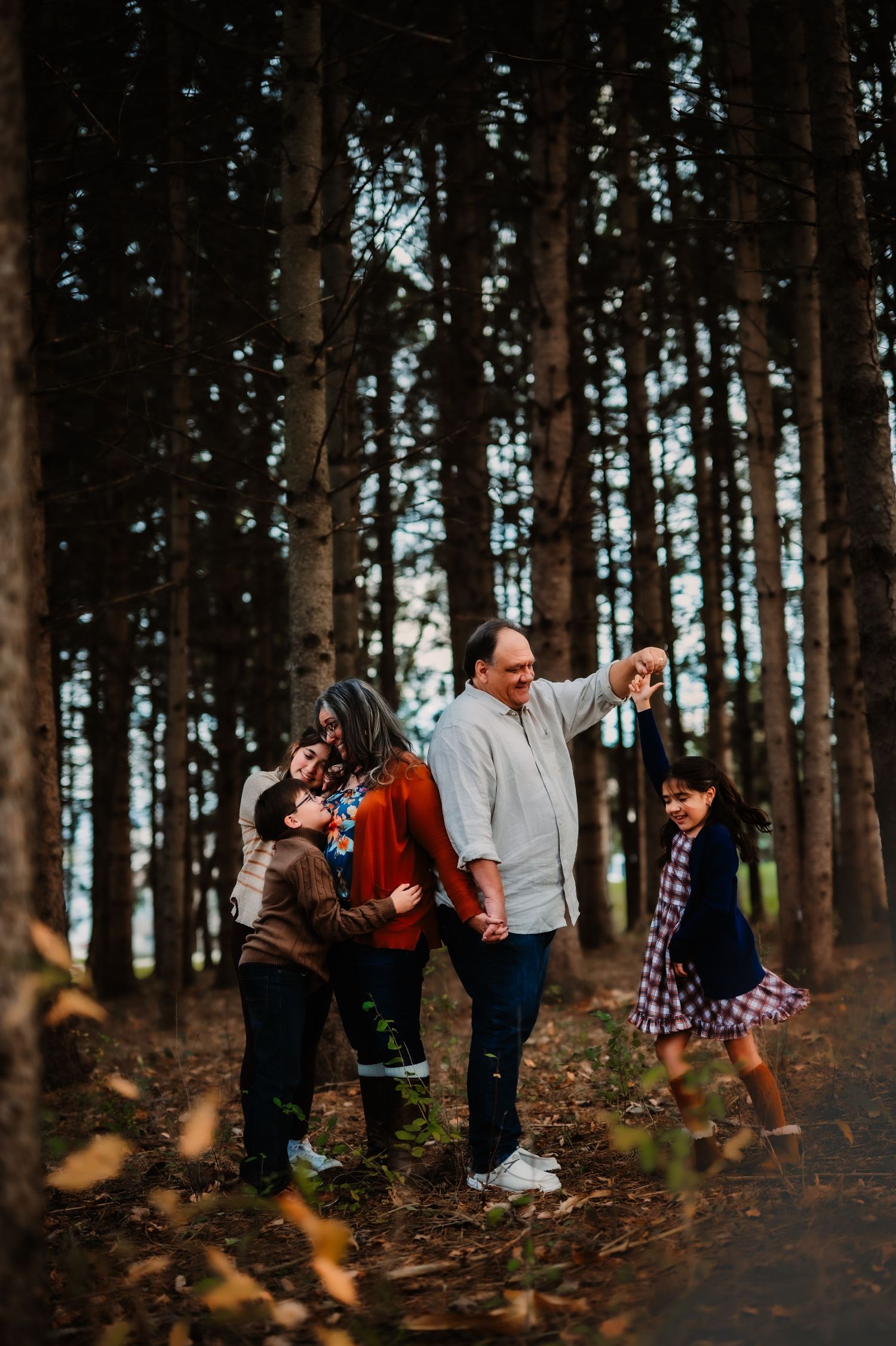 Family of four enjoying a walk through tall pine forest, parents spinning young daughter while older child watches.